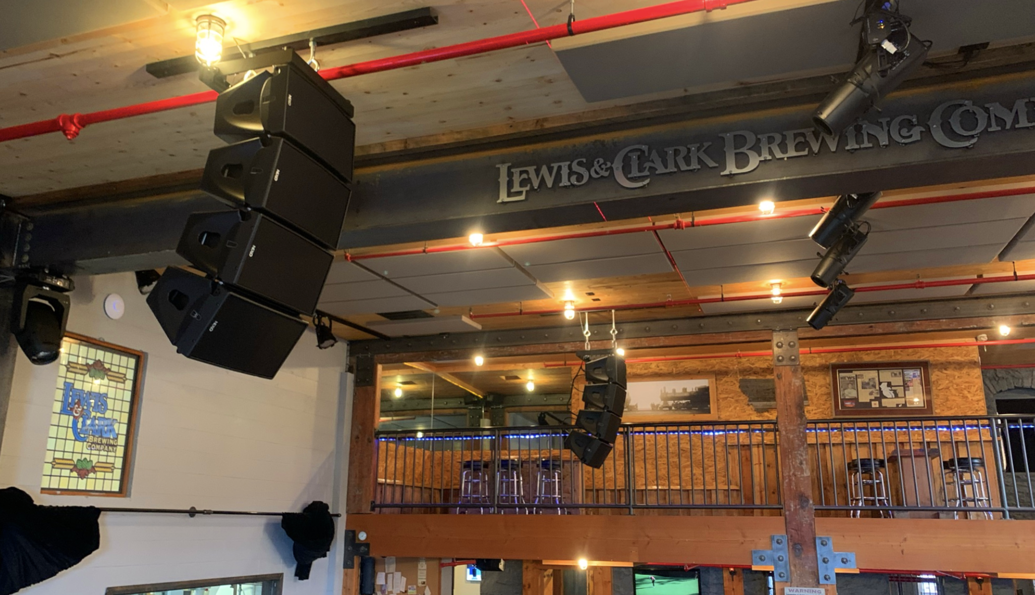 Interior of a brewery with a stage area featuring large black speakers, wooden beams, a railing, and framed signs on the wall, including a Lewis & Clark Brewing sign.