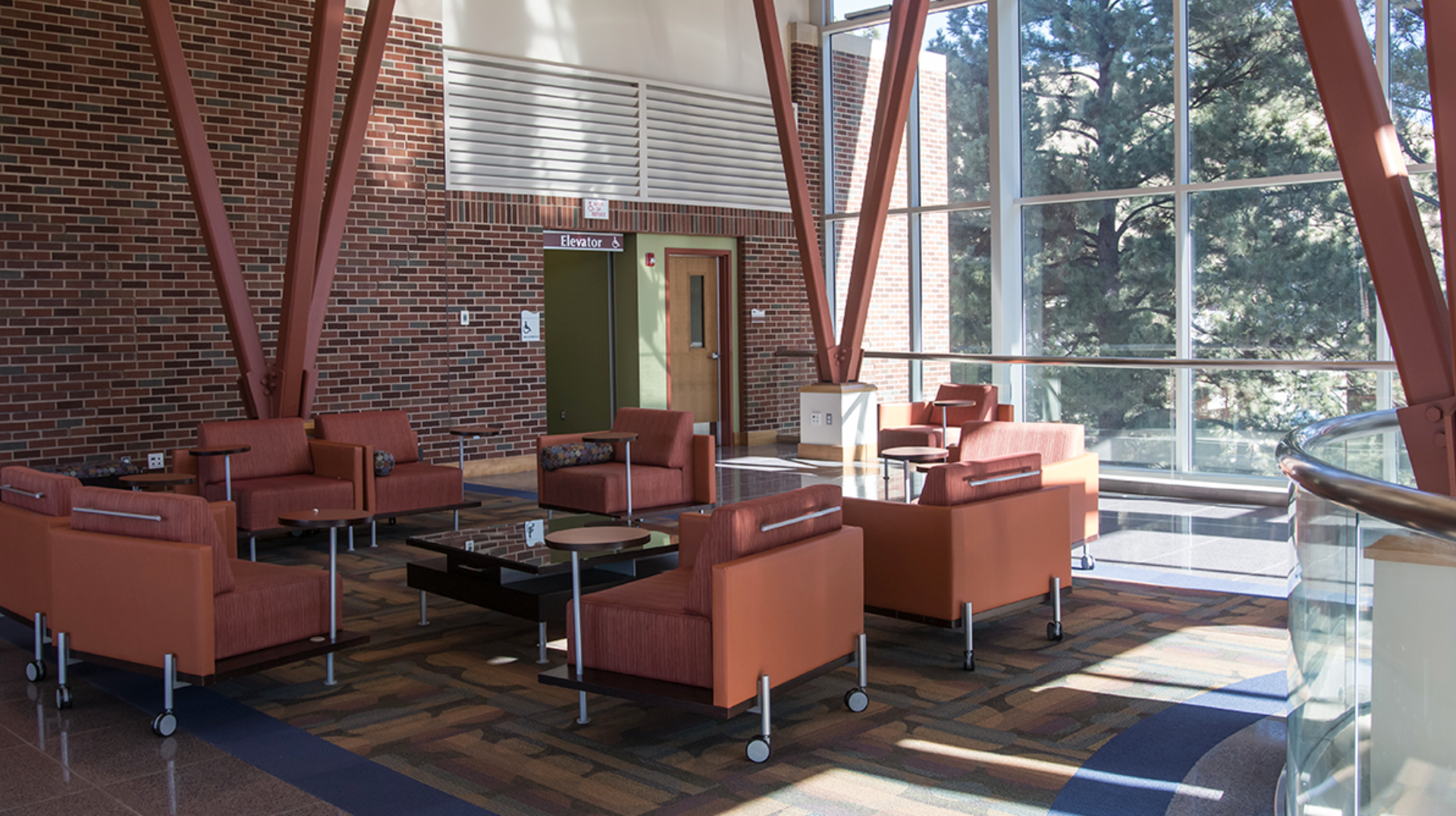 Modern indoor lounge area with orange upholstered chairs and sofas, glass walls showing trees outside, and a brick wall.