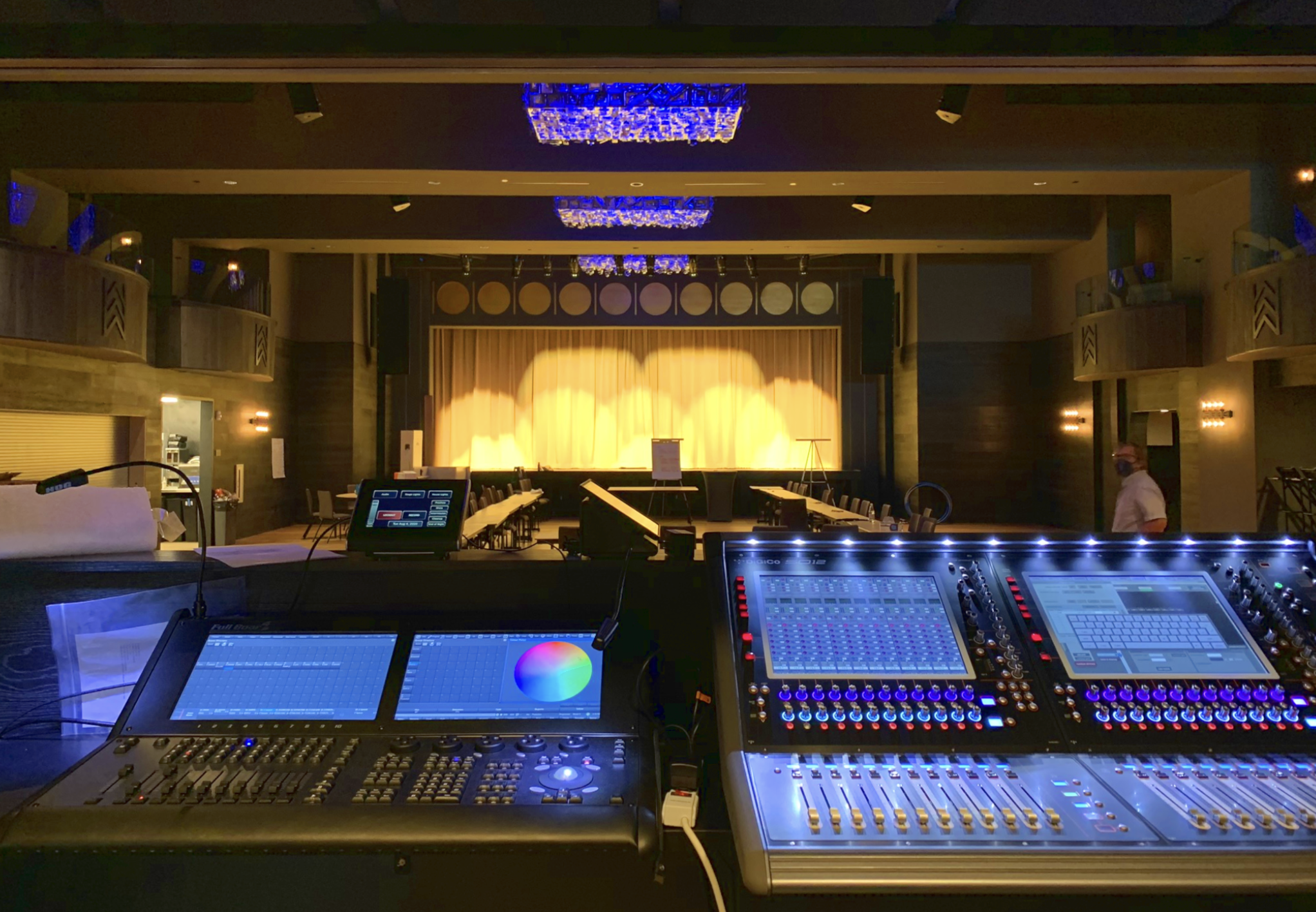 Empty stage with yellow curtains and lighting effects, viewed from behind a sound mixing console with electronic equipment and controls in the foreground.