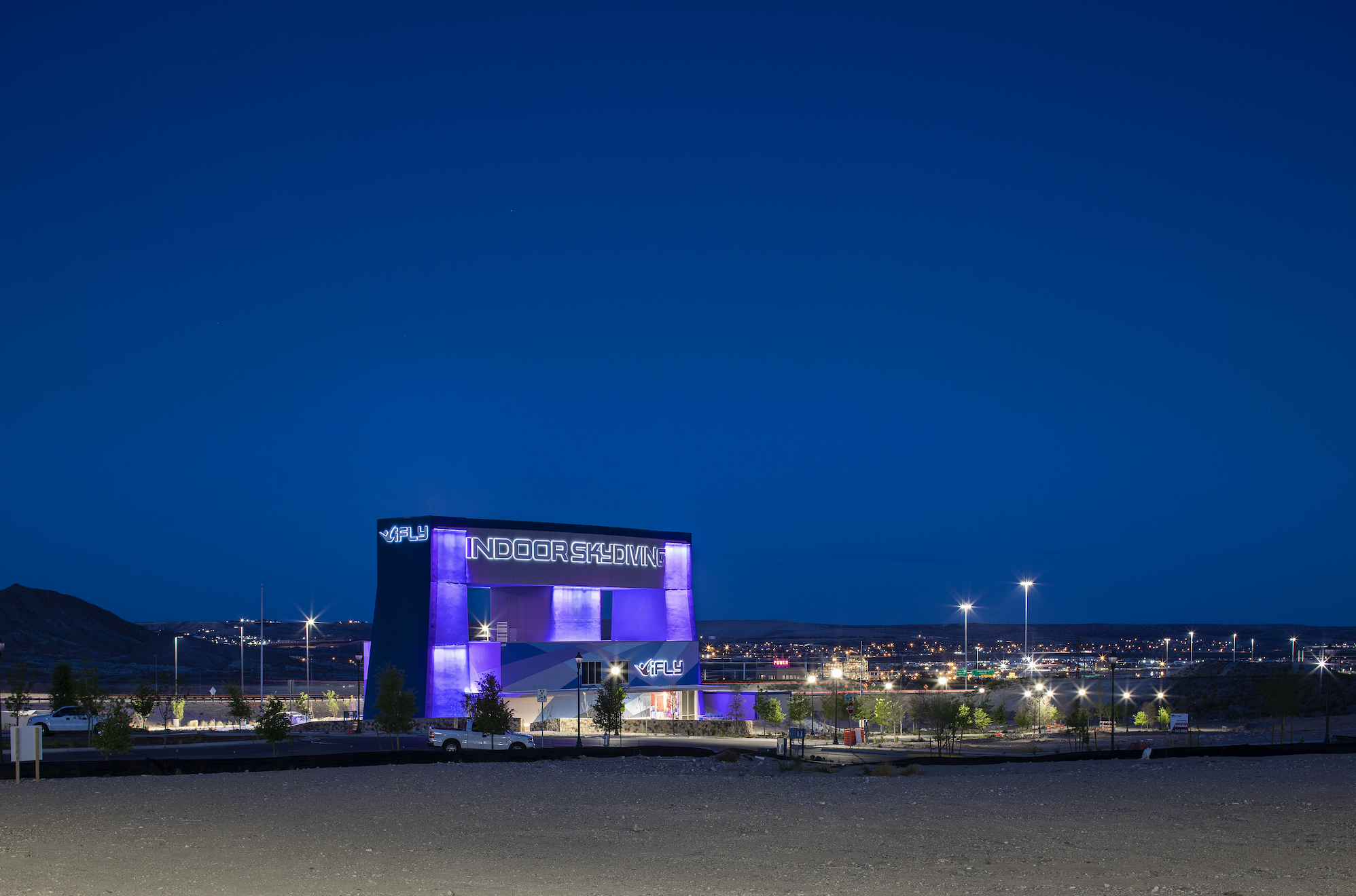 Nighttime view of an indoor skydiving facility with purple lighting, illuminated signs, and surrounding parking lot with streetlights and trees.
