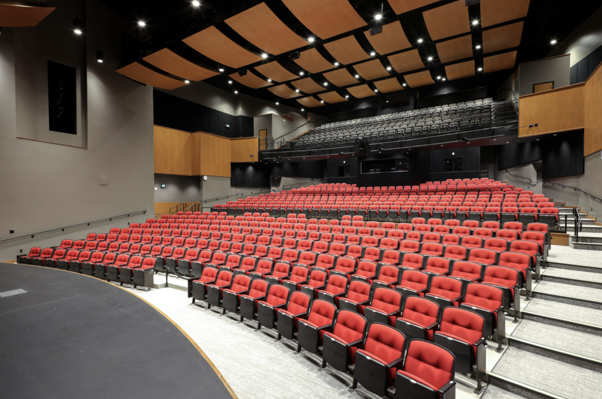 Interior view of a modern theater auditorium with red and black seats, a stage on the left, and acoustic panels on the ceiling.