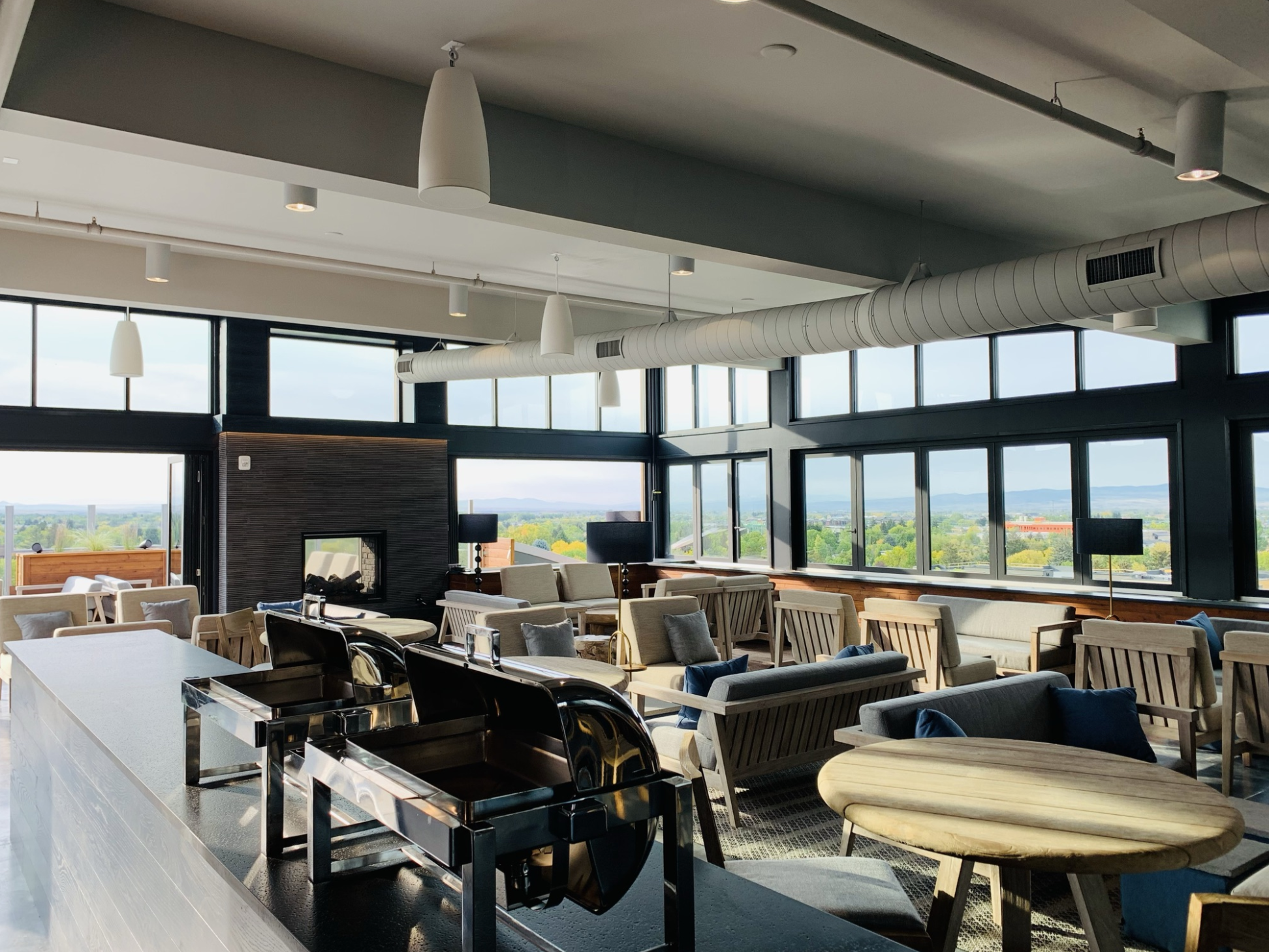 Modern lounge with large windows, beige and gray seating, round wooden tables, and black lamps, overlooking a cityscape and green landscape.