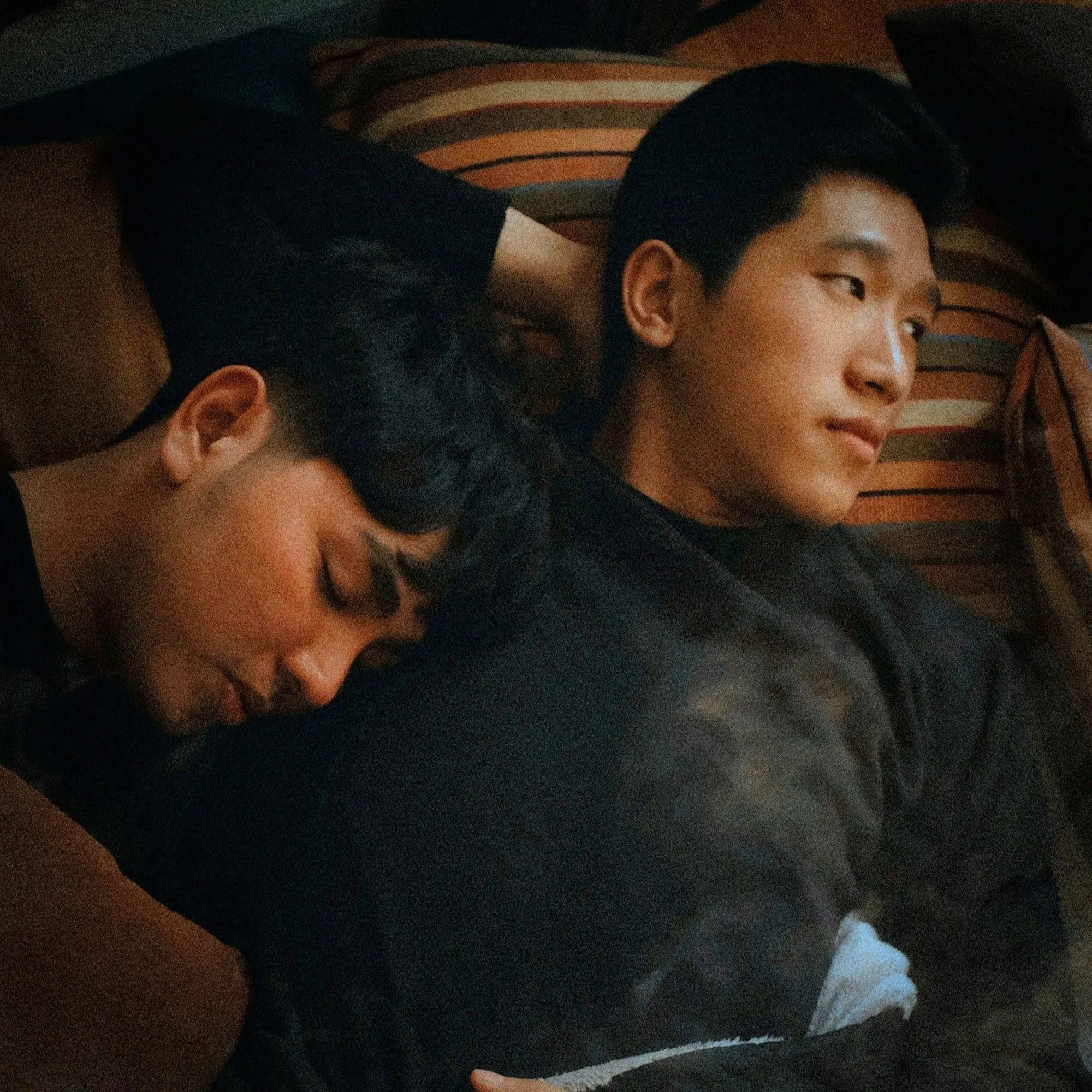 A young person with short dark hair and a slight smile lying on a striped pillow with their head turned to the side in a dark room.