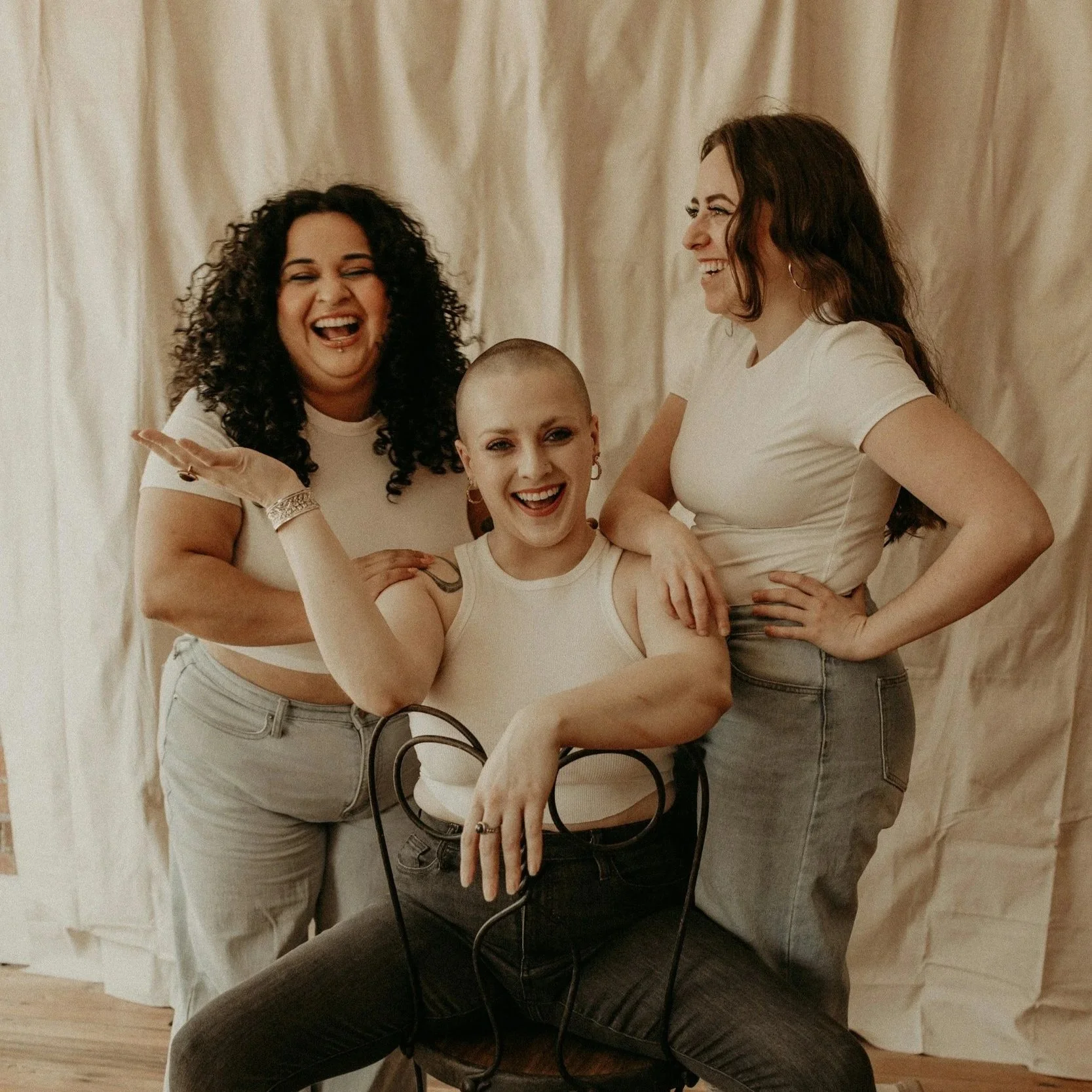 Three women posing together, one sitting on a chair, all smiling and laughing, with a beige curtain background.