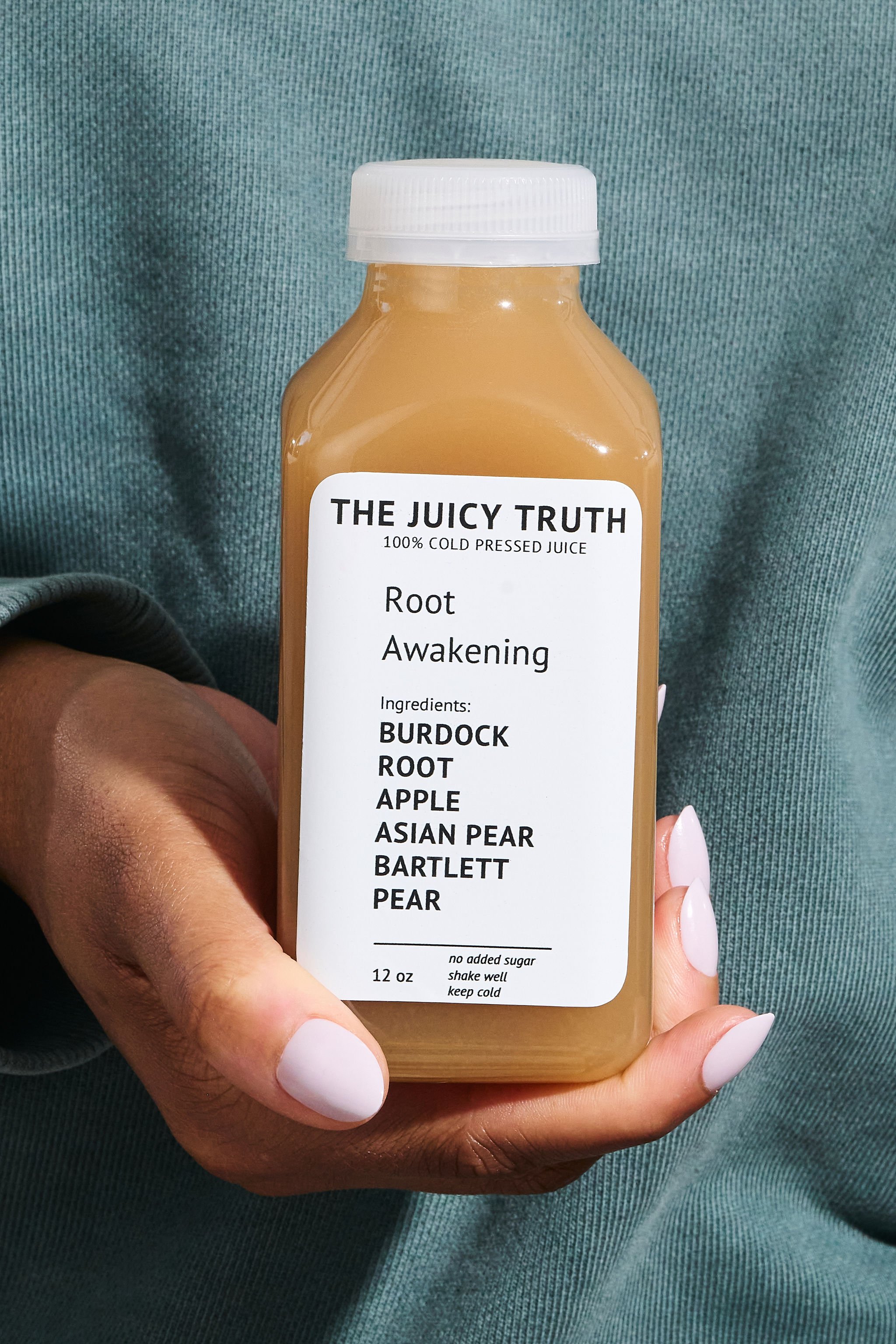 A hand holding a bottle of cold pressed juice labeled 'The Juicy Truth' with ingredients including burdock root, apple, Asian pear, Bartlett pear, and the word 'Awakening' on the label.