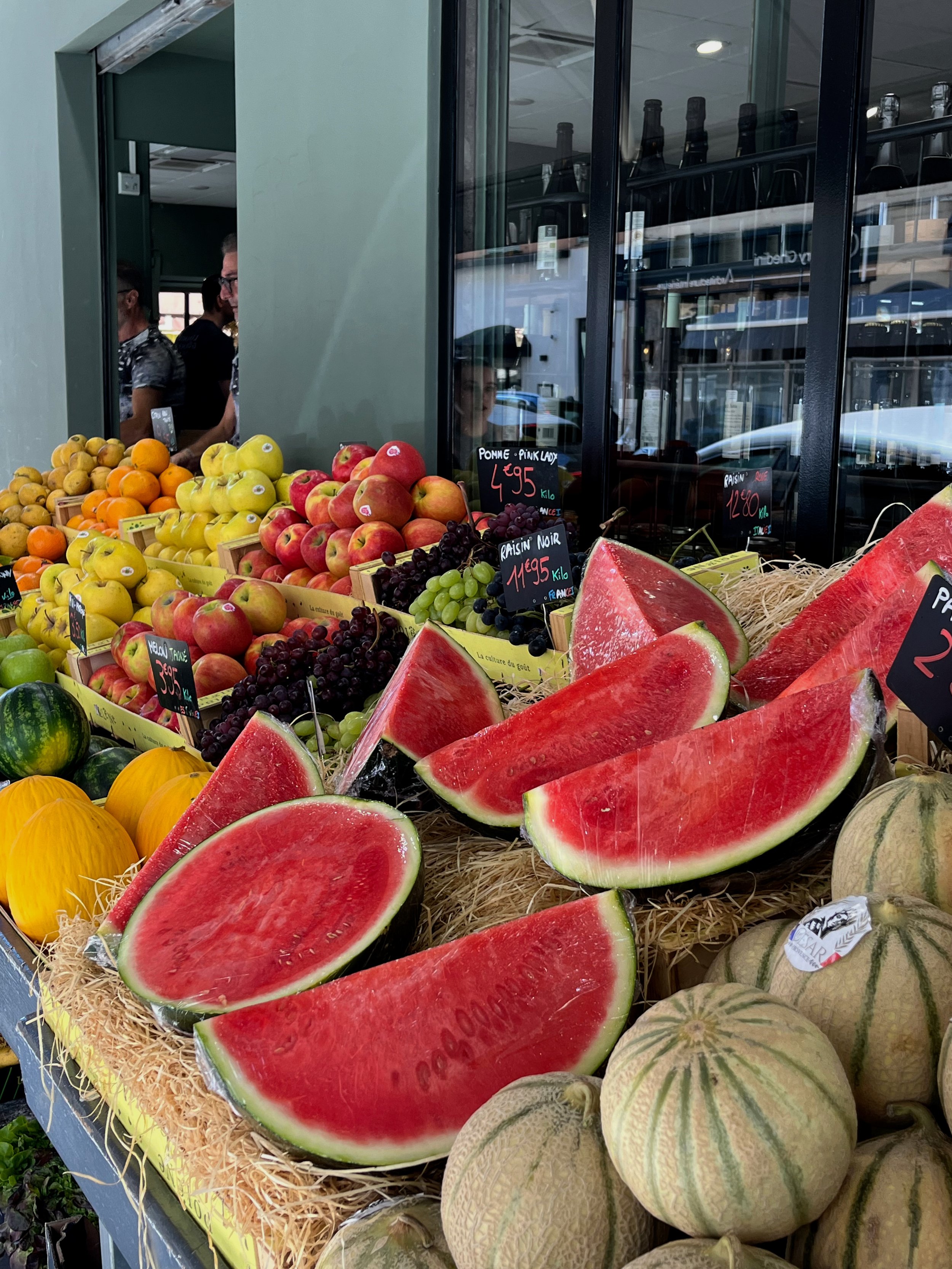 Display of fresh sliced watermelon and whole melons at a fruit stand, with other fruits like apples, grapes, and persimmons in the background.