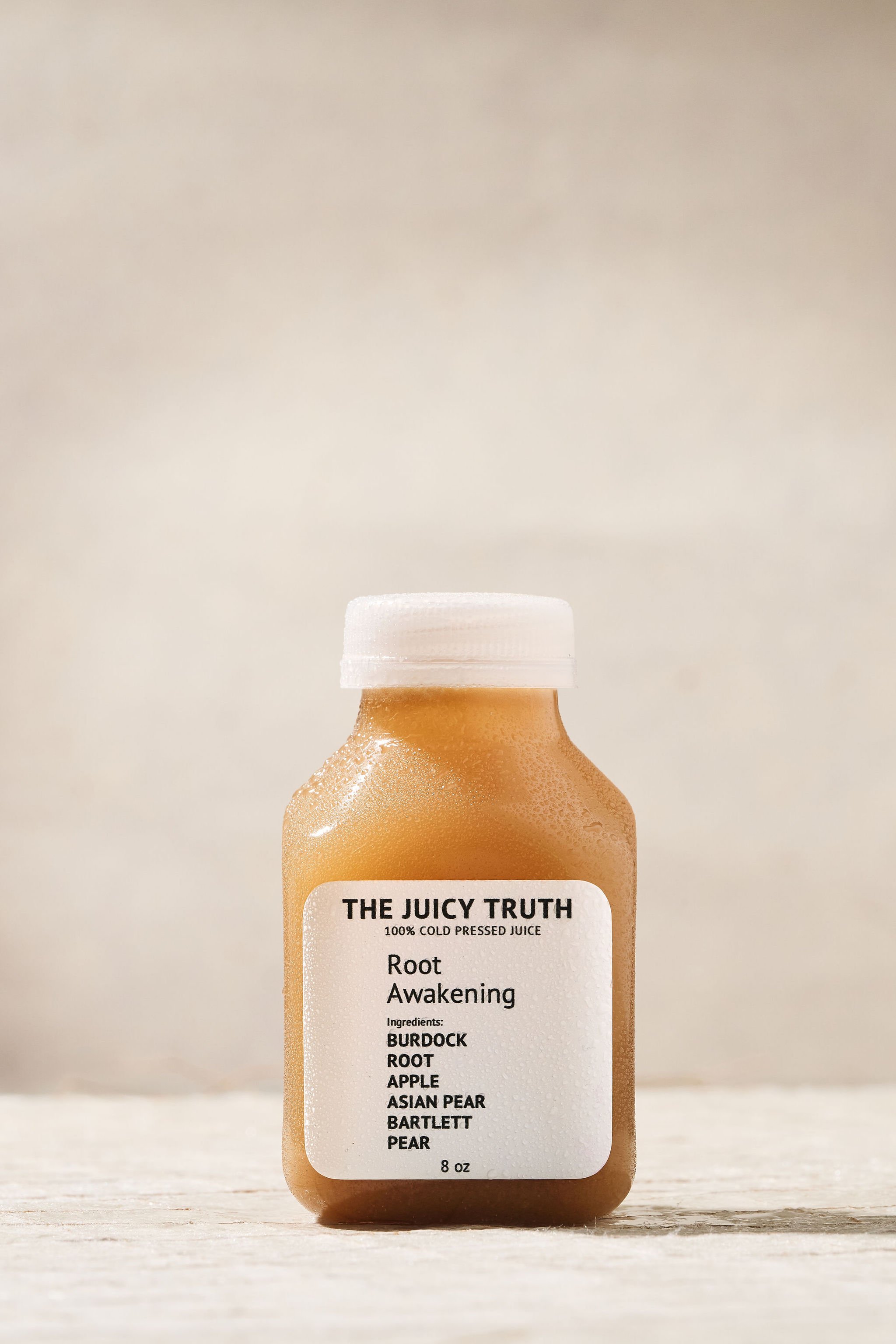 A small bottle of cold pressed juice labeled 'The Juicy Truth' with ingredients including burdock root, apple, Asian pear, Bartlett pear, and water. The bottle is covered in condensation, and has a white cap, placed on a light wooden surface against