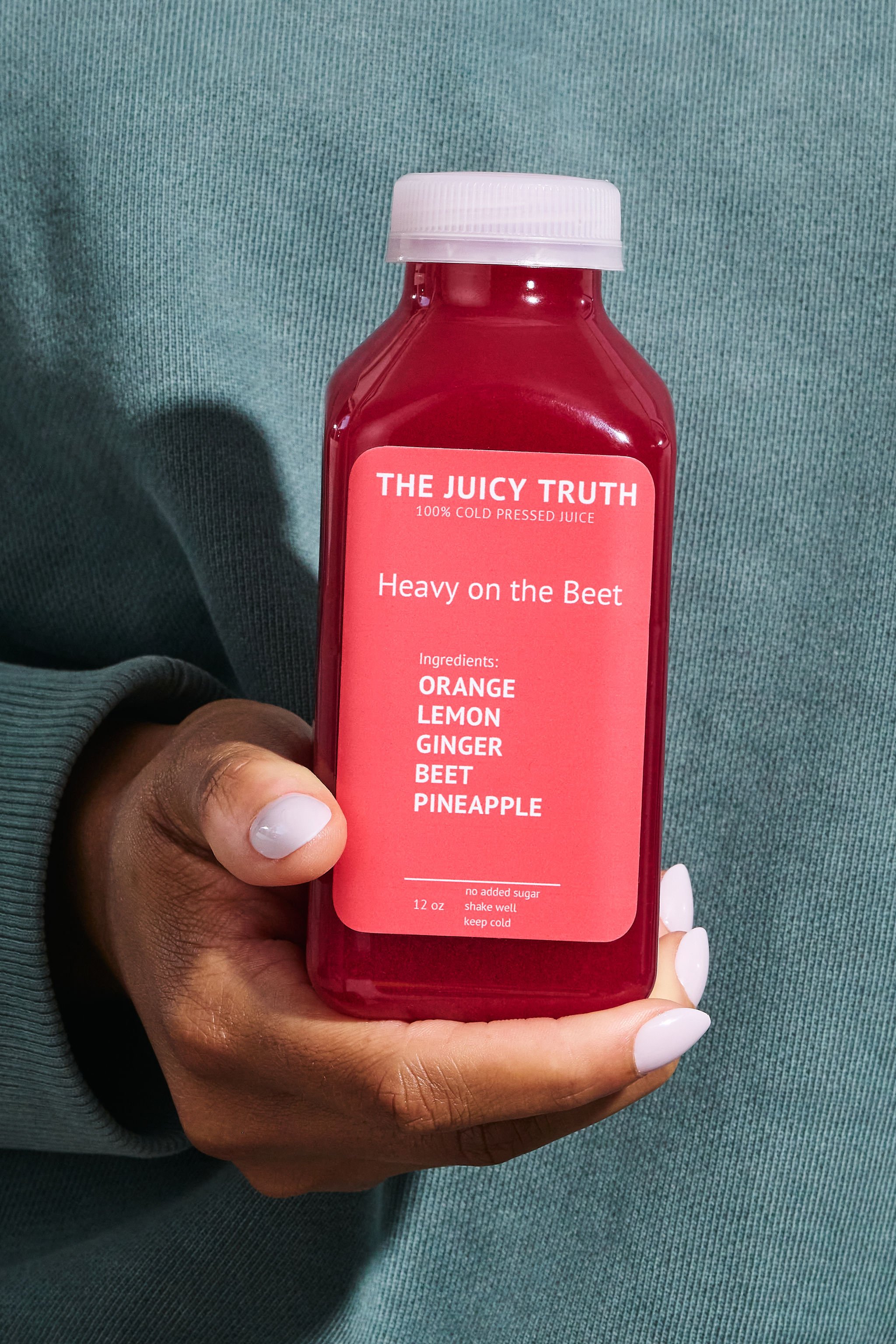 A hand holding a red bottle of juice labeled 'The Juicy Truth' with ingredients listed as orange, lemon, ginger, beet, and pineapple.