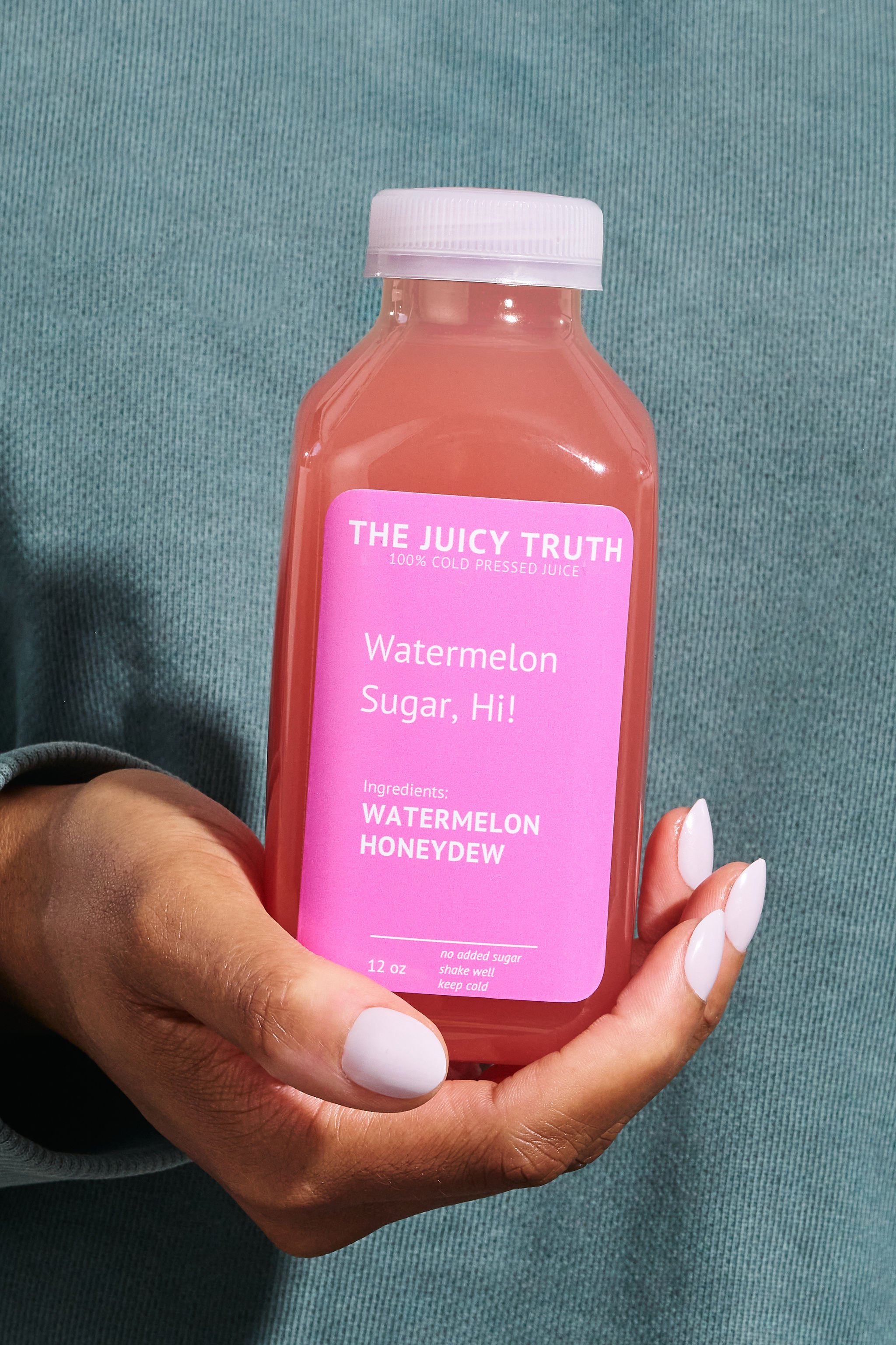 A hand holding a pink bottle of watermelon juice labeled 'The Juicy Truth', with ingredients listed as watermelon and honeydew, against a teal fabric background.