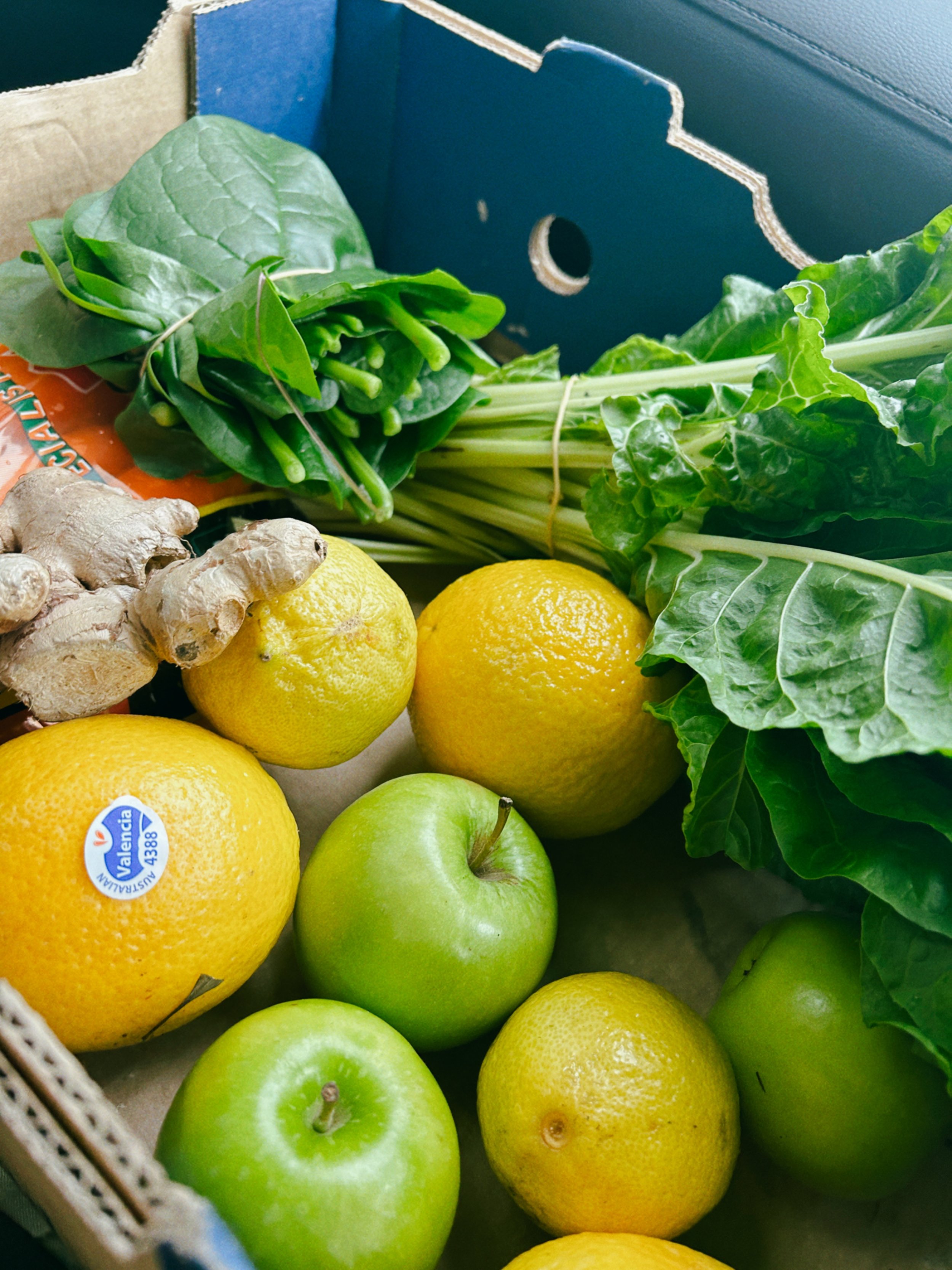 Fresh lemons, green apples, ginger, and leafy greens in a cardboard box.