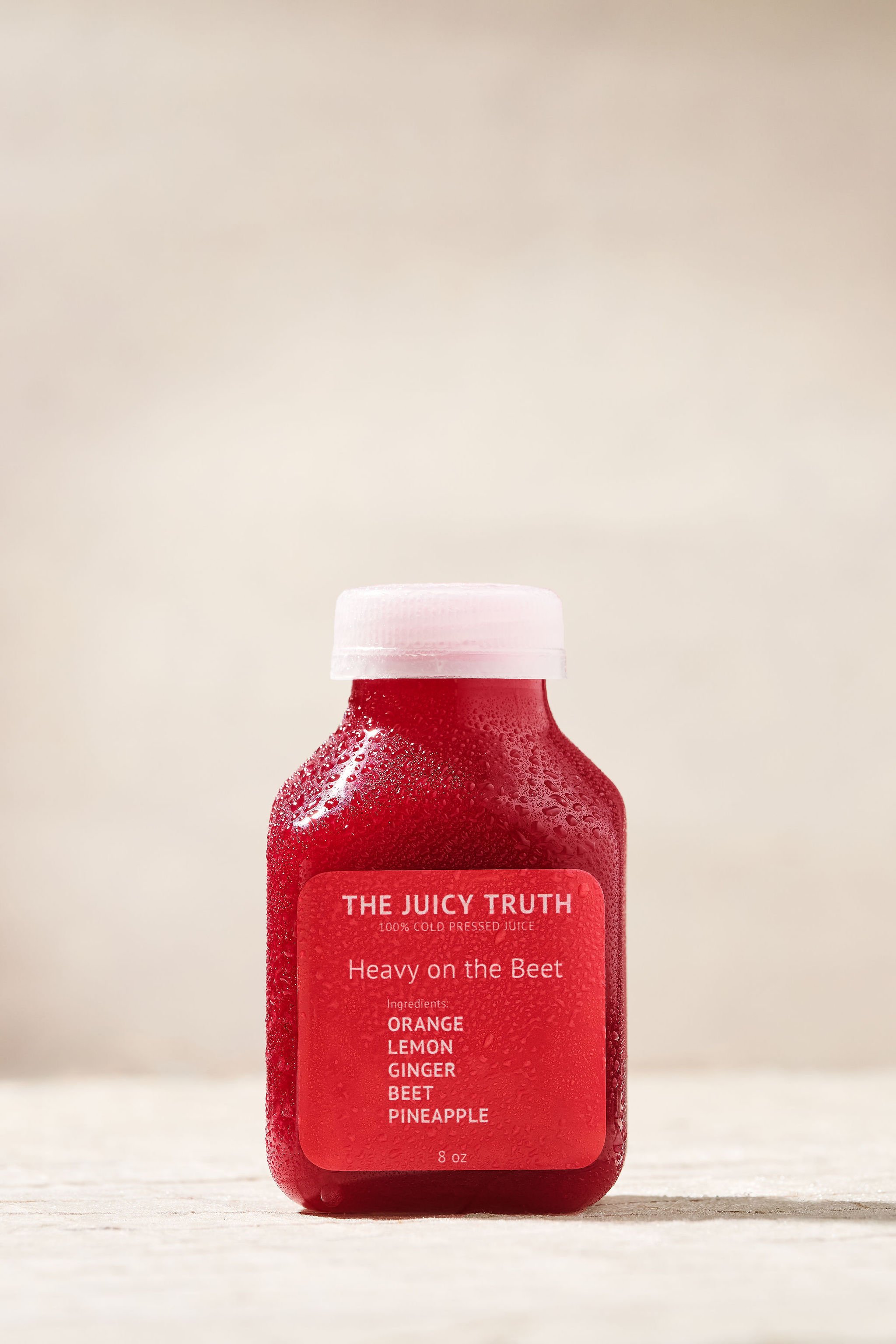 Red bottle of cold pressed juice labeled "Heavy on the Beet" with ingredients including orange, lemon, ginger, beet, and pineapple placed on a light-colored surface with condensation on the bottle.