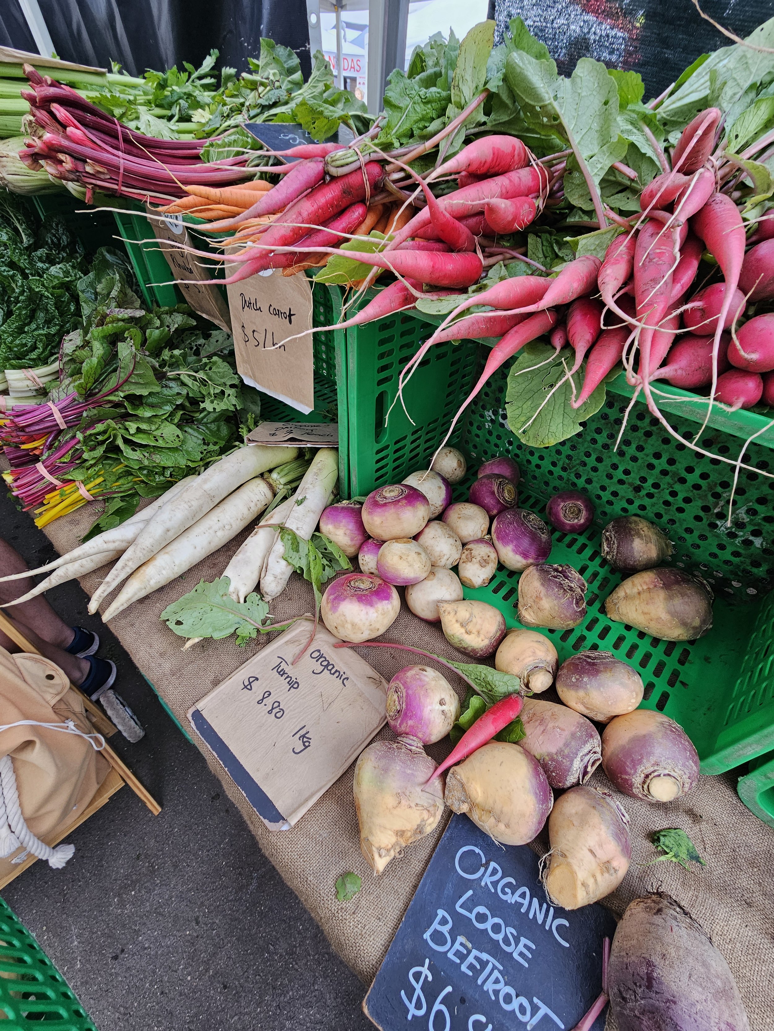 Fresh organic carrots, turnips, and beets displayed at a farmer's market. Signs show prices for each type of vegetable.
