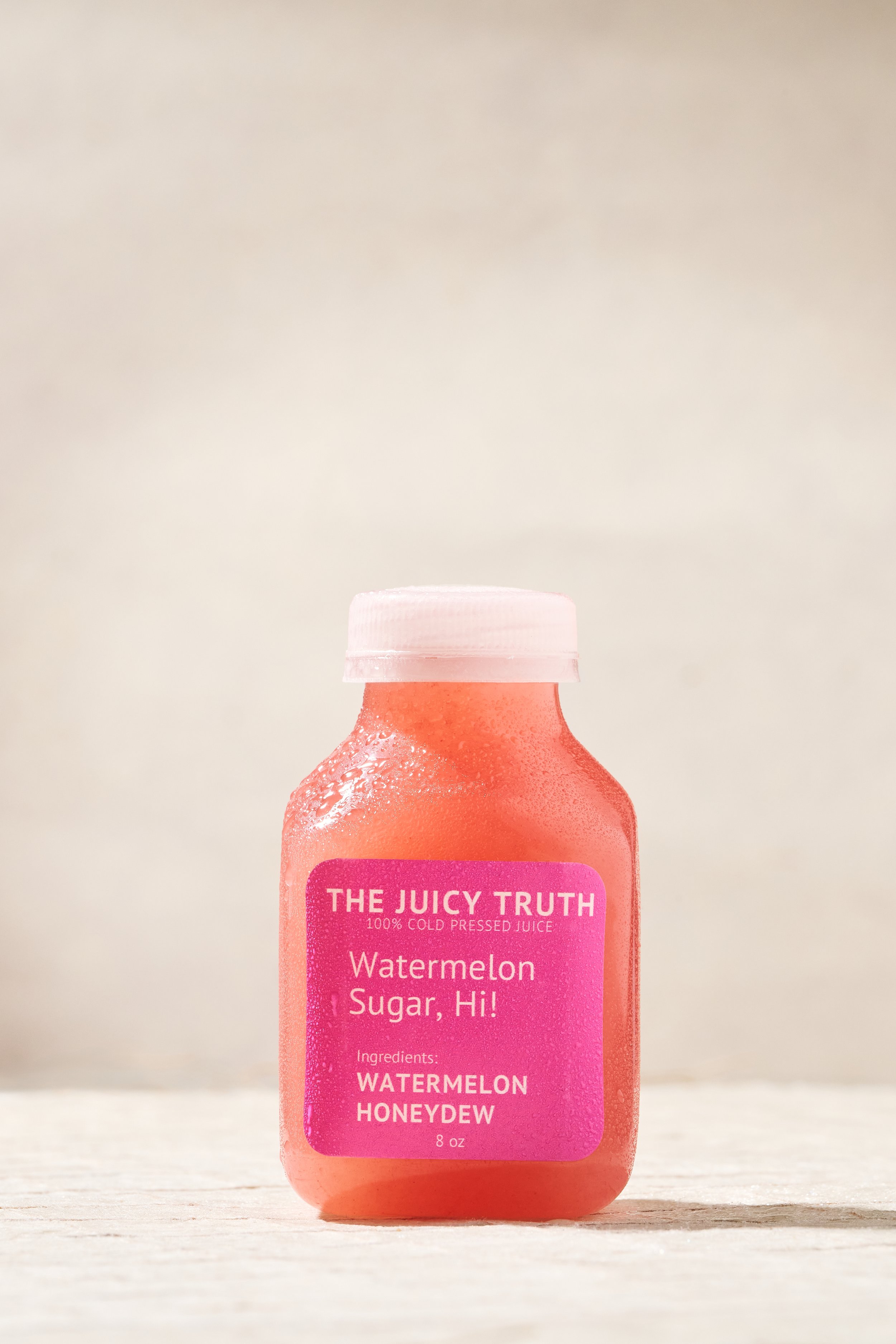 A small bottle of pink-colored watermelon honeydew juice labeled 'THE JUICY TRUTH 100% COLD PRESSED JUICE Watermelon Sugar, Hi! Ingredients: WATERMELON HONEYDEW 8 oz' with condensation on the bottle, placed on a light wooden surface against a plain l