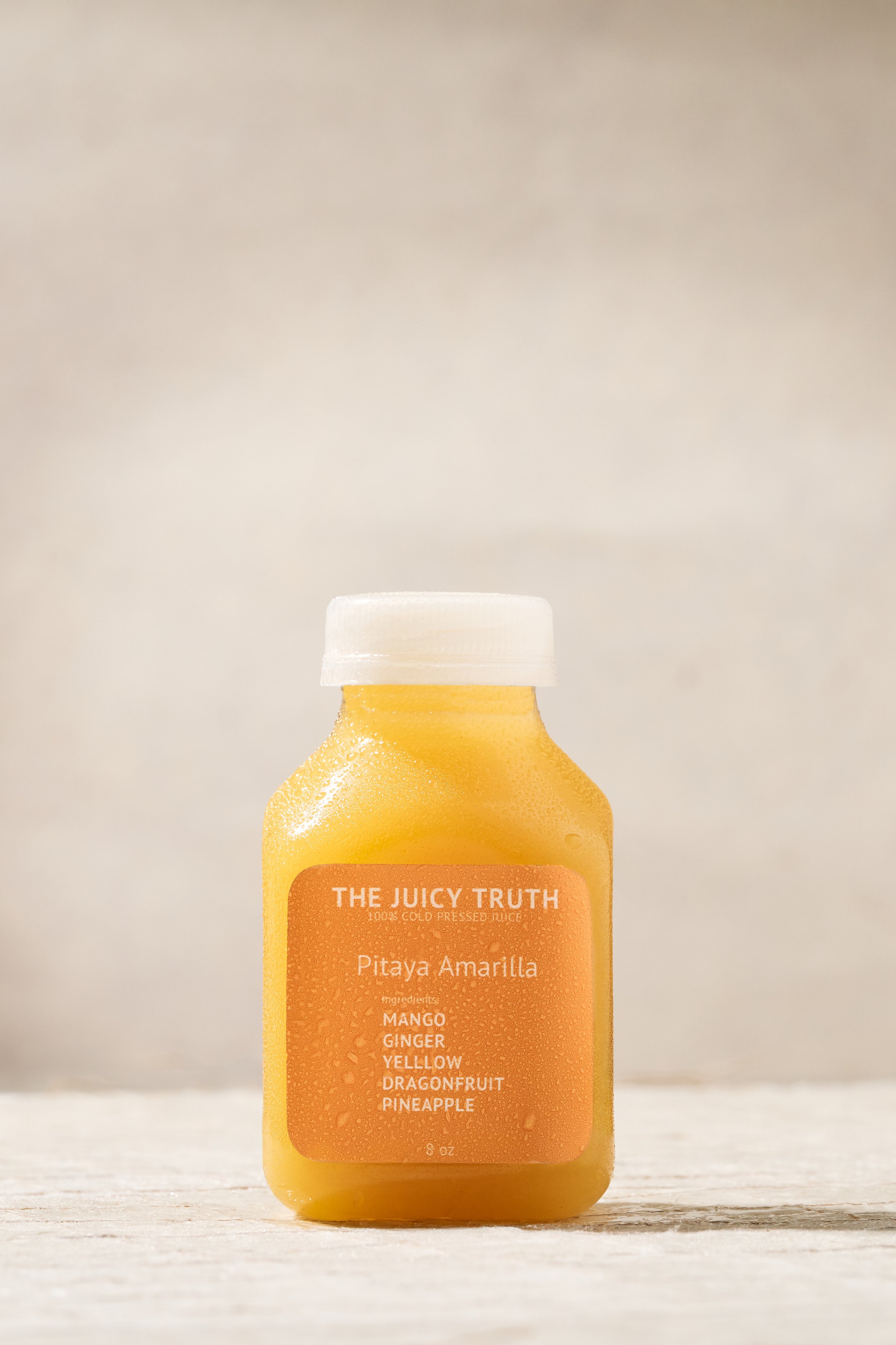 Bottle of yellow juice labeled 'The Juicy Truth Pitaya Amarilla' with ingredients including mango, ginger, yellow, dragonfruit, and pineapple, sitting on a wooden surface with a light background.