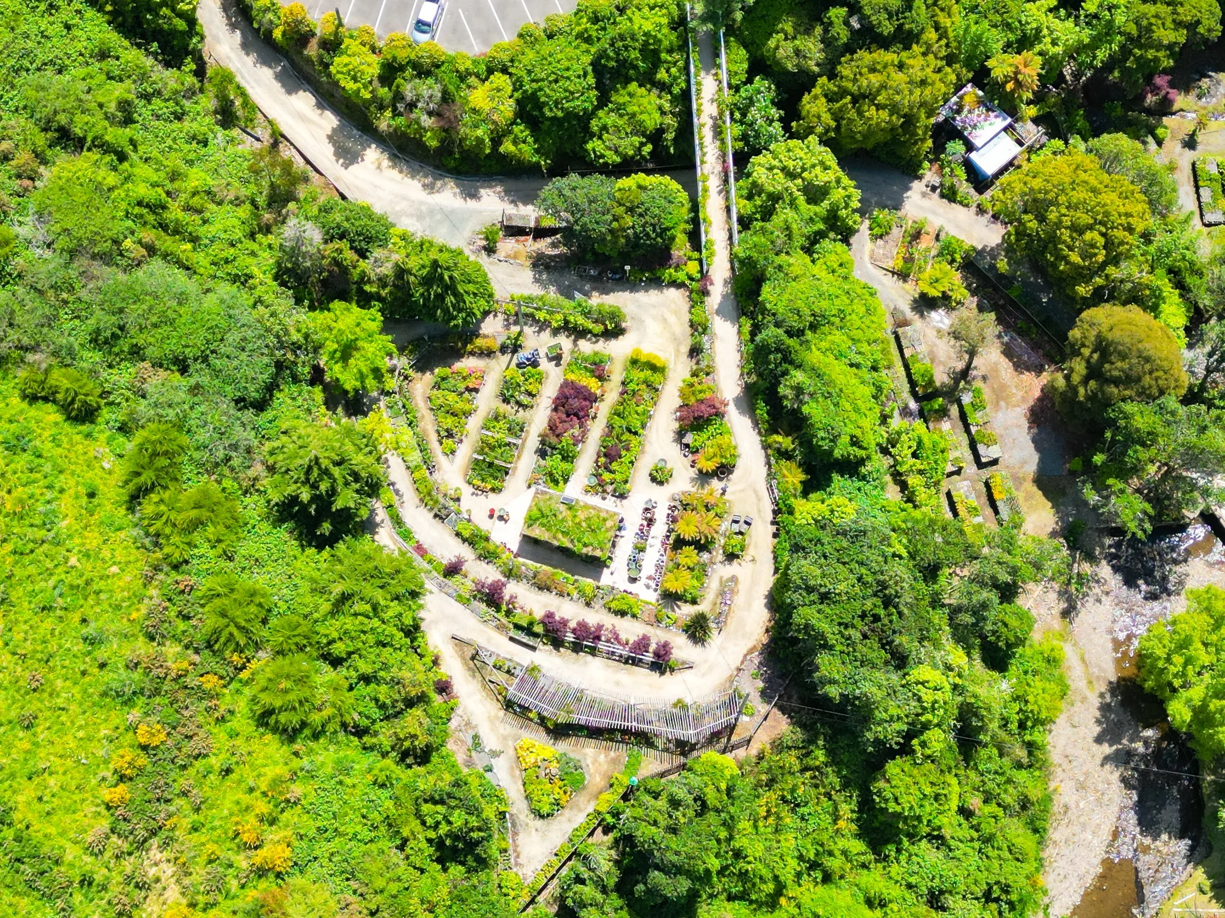 An aerial view of a lush, green landscape with a winding dirt road leading to a small garden and parking area. The area is surrounded by dense trees and vegetation, with a few buildings visible in the distance.