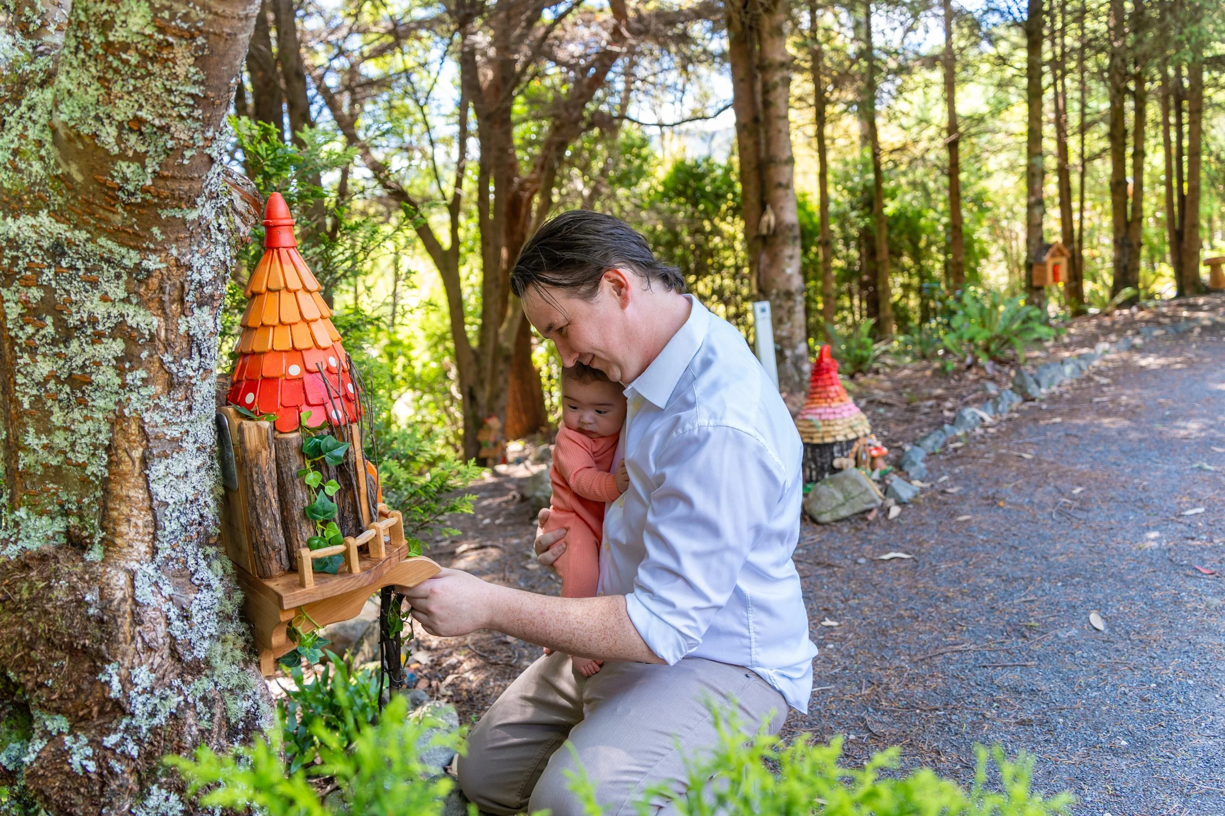 A man kneeling on the ground in a wooded area, holding a young child, with decorative birdhouses mounted on a tree and along the trail in the background.