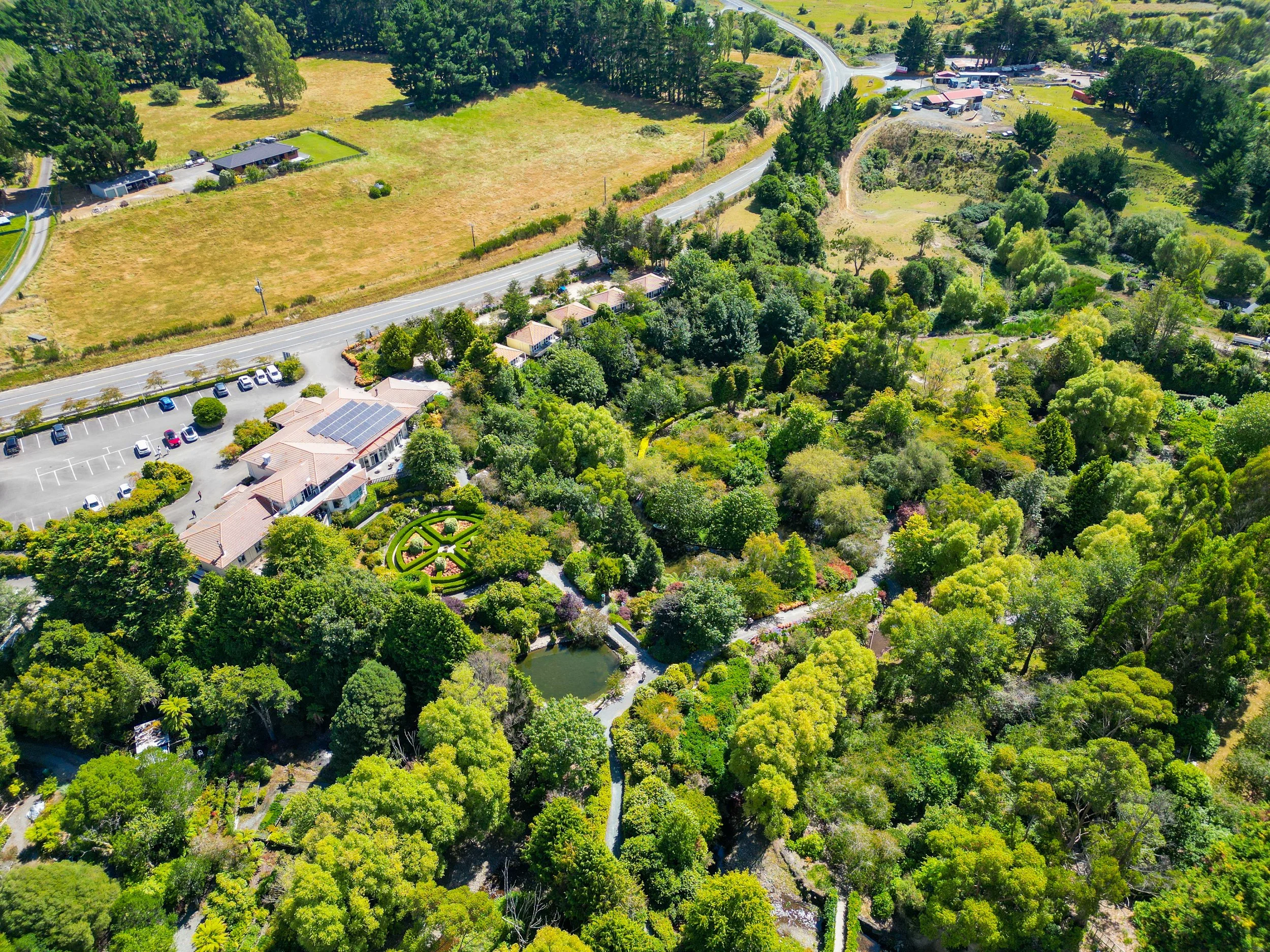 Aerial view of a lush, green community with a large, well-maintained garden, surrounding trees, a pond, a parking lot, and roads.