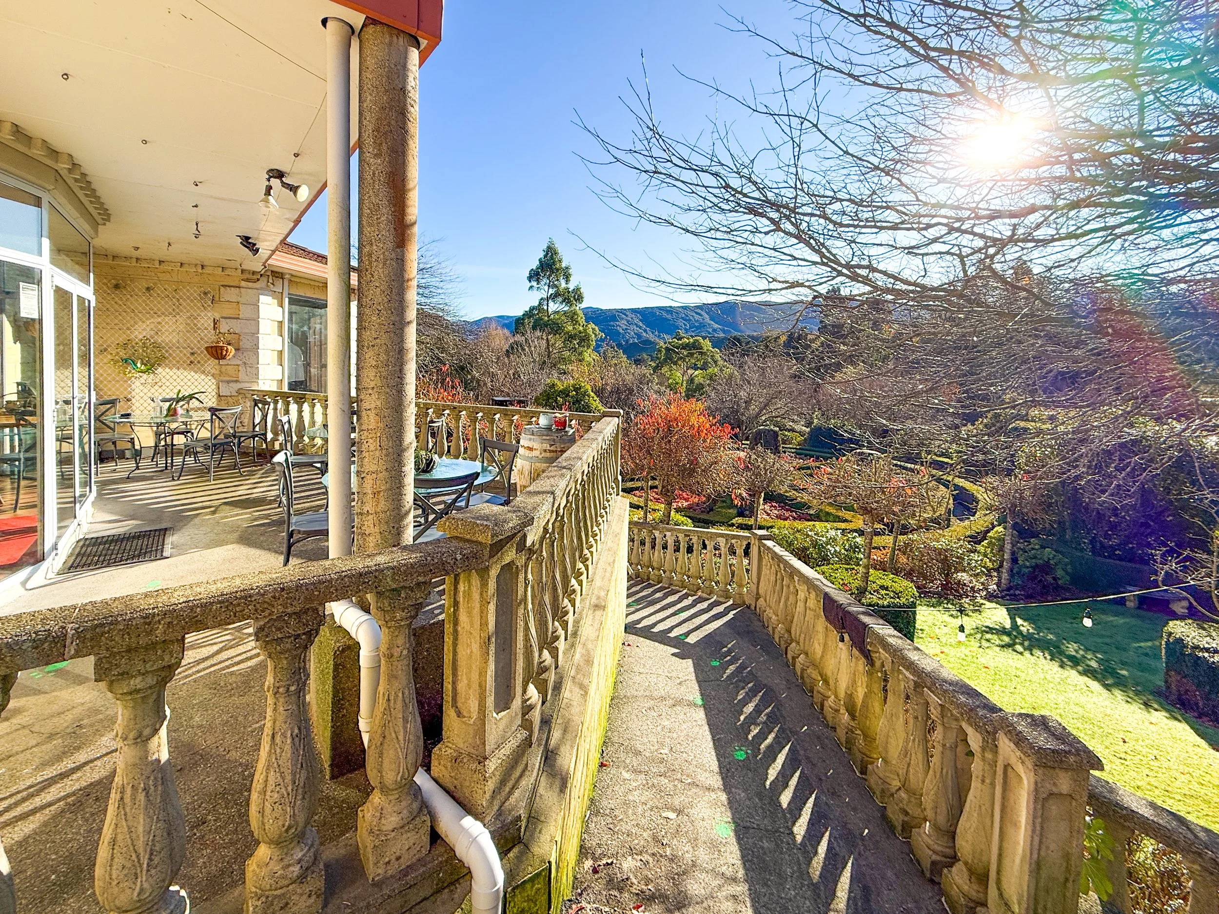A balcony with outdoor seating overlooking a garden with trees and mountains in the background on a sunny day.