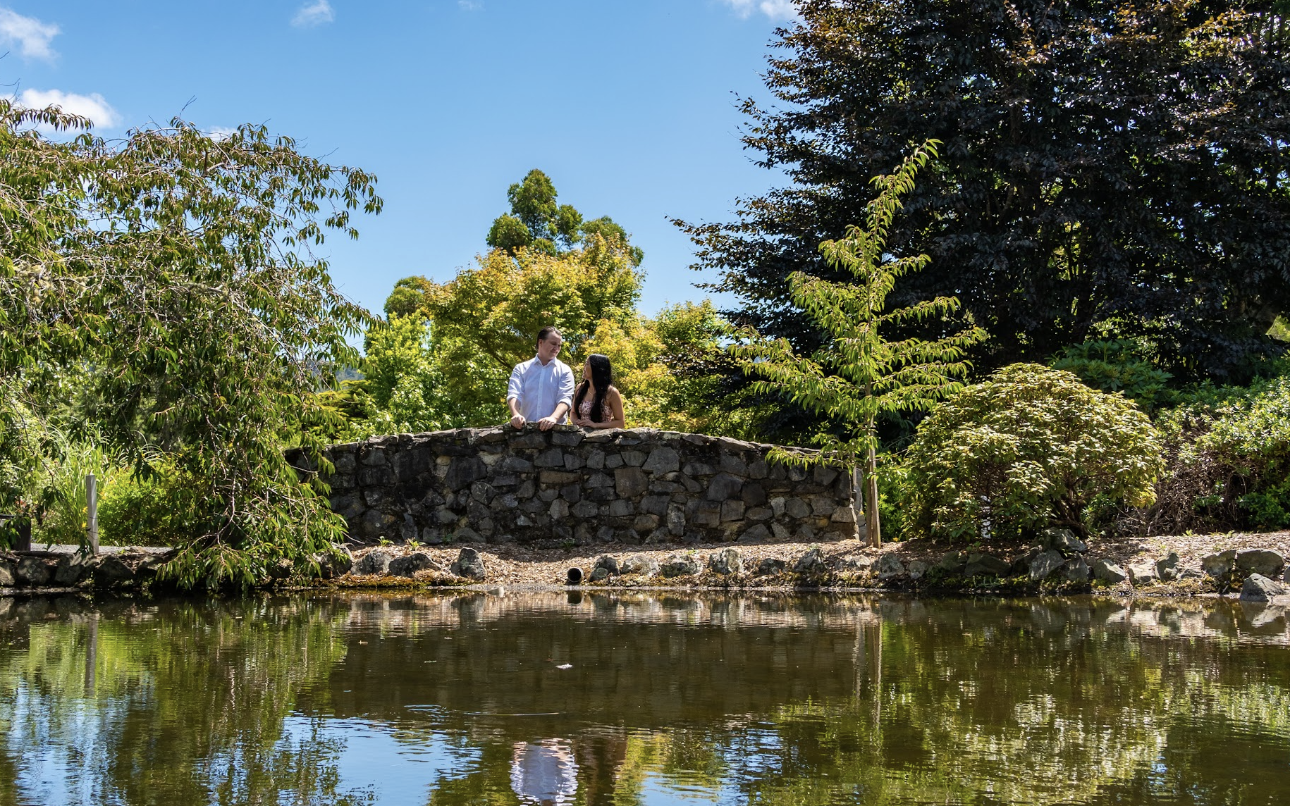 A young man and woman stand on a stone bridge over a pond, surrounded by lush green trees under a clear blue sky.