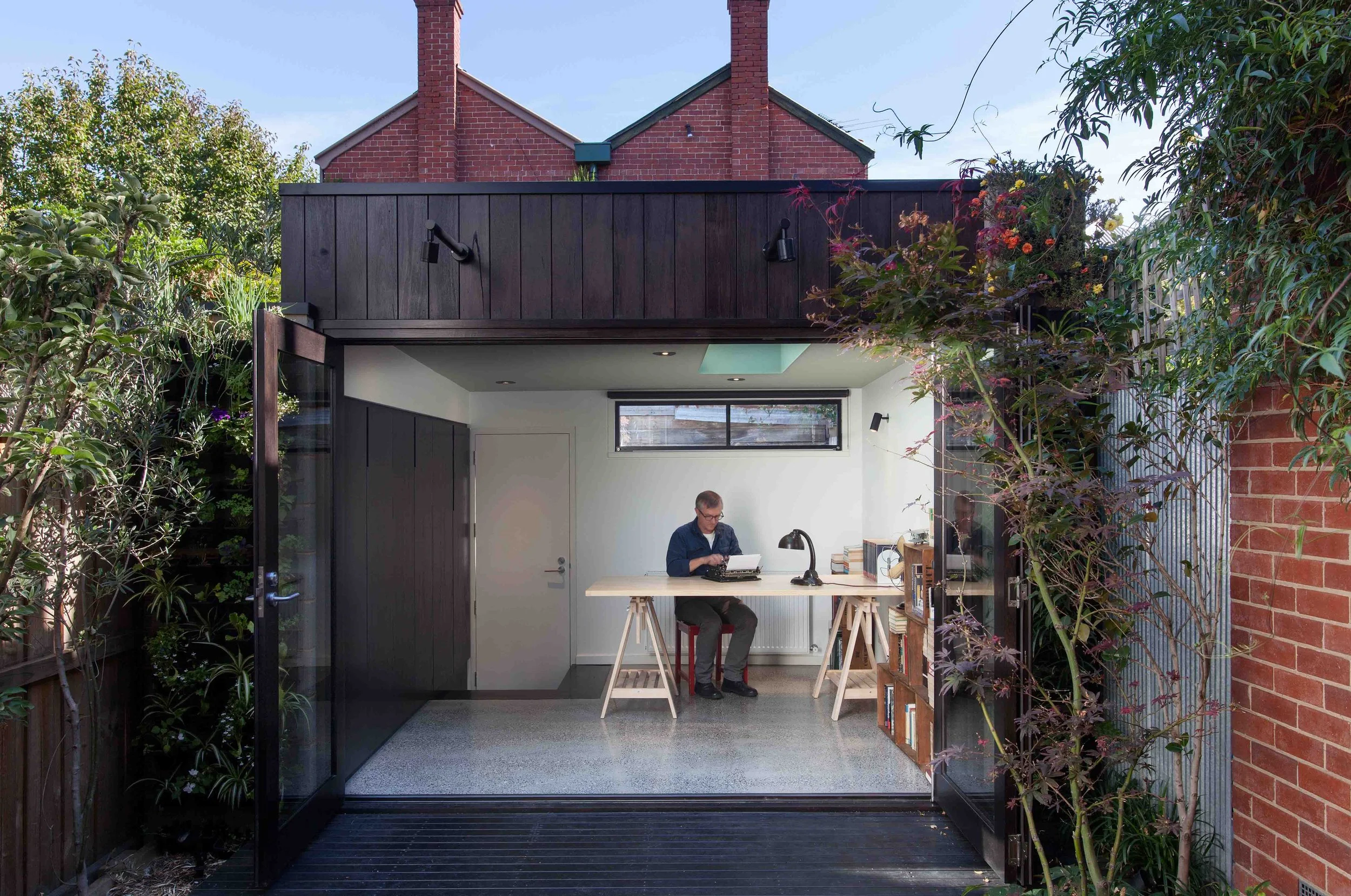 Compact garden studio with fully opened glass doors connecting the workspace to the surrounding garden and timber deck.