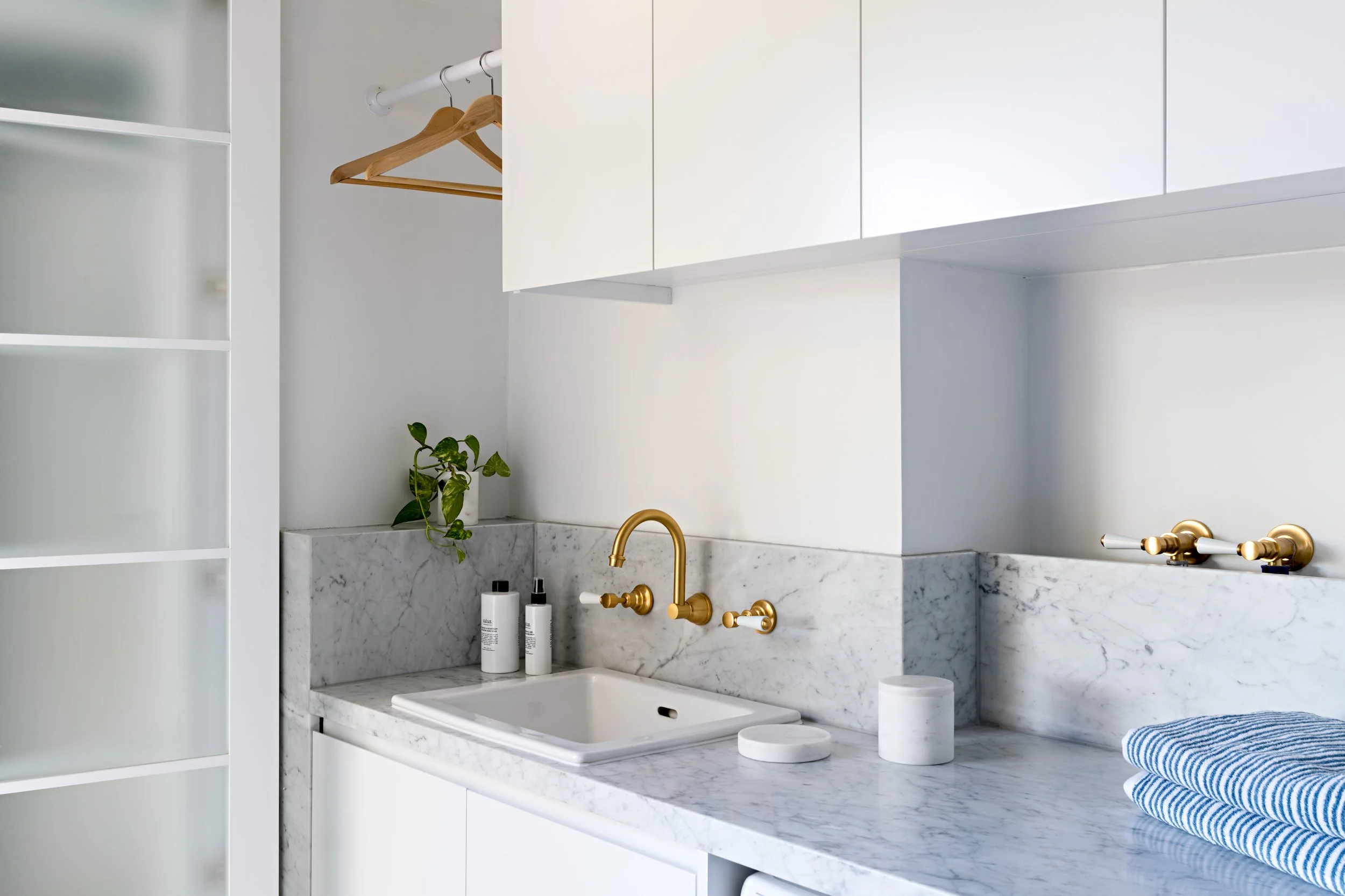 White laundry with marble benchtop and splashback, overhead cabinetry and brass tapware.