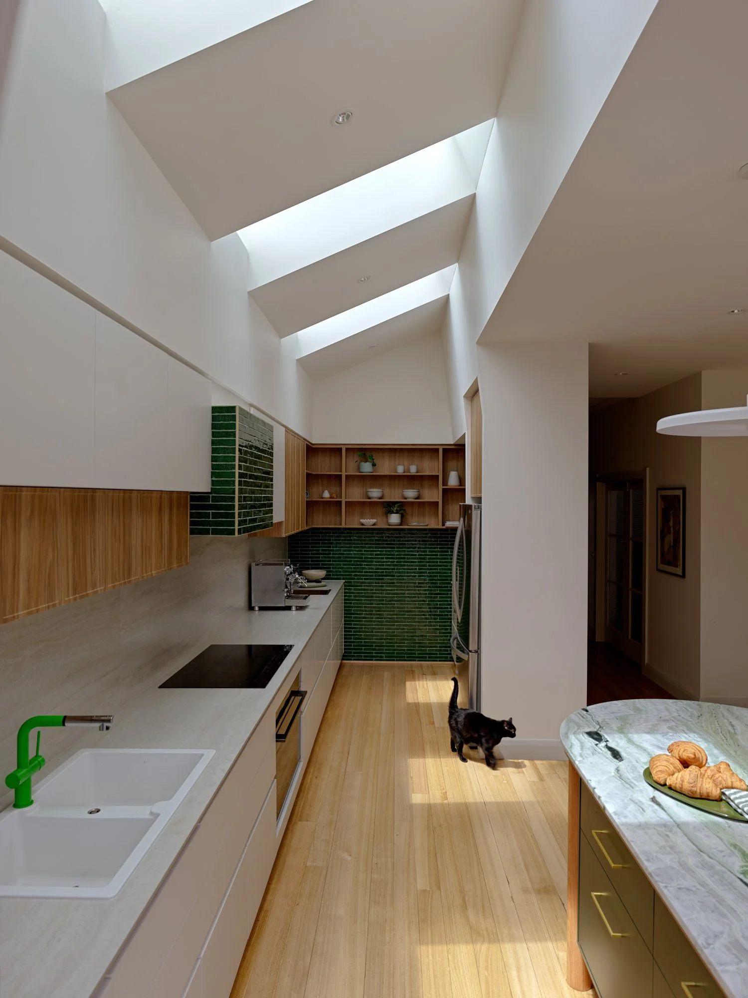 View along the Northcote Extension kitchen showing the long benchtop, green tiled wall, open shelving and series of skylights above.