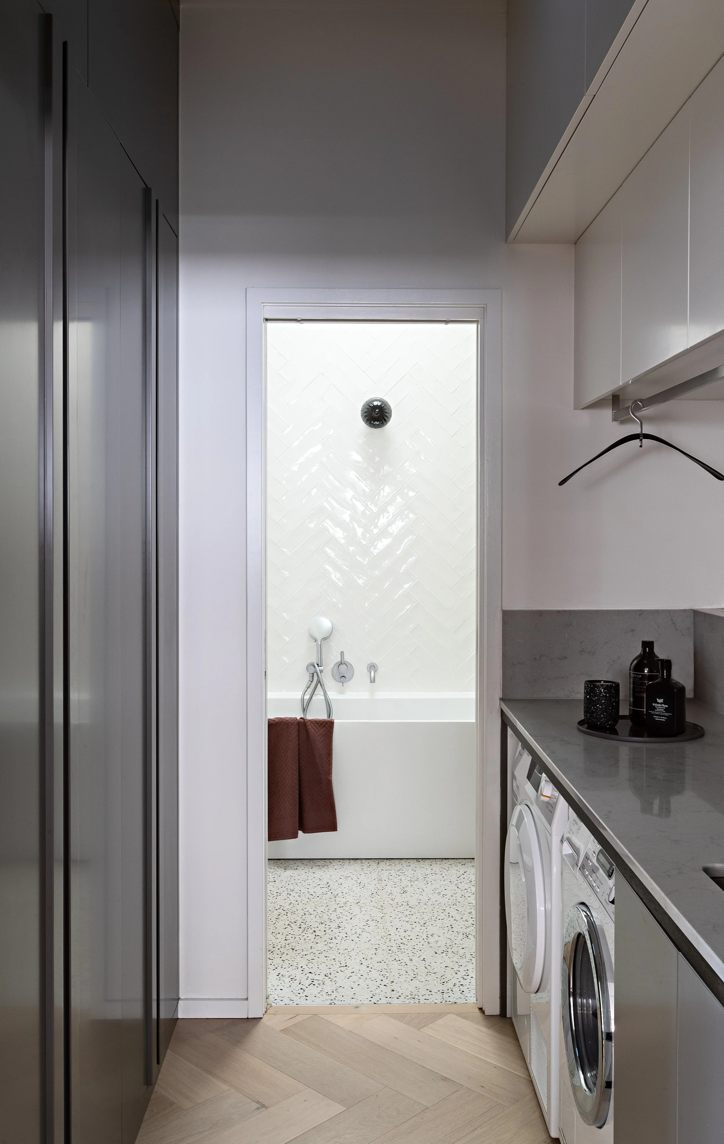 Compact laundry with dark cabinetry leading through to a light-filled bathroom at Brighton East House.