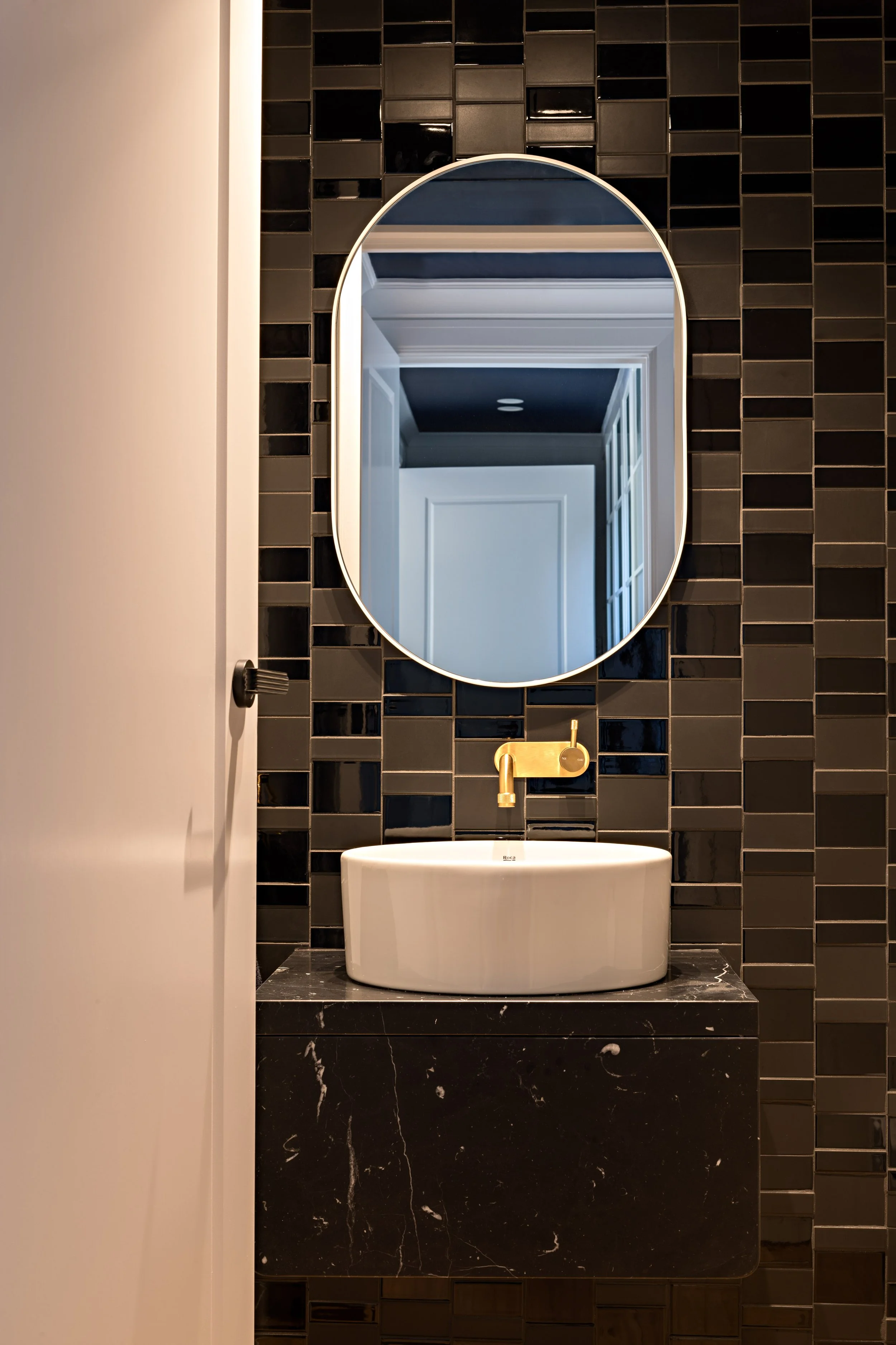 Powder room with black tiled walls, black marble vanity, oval mirror and brass tapware.