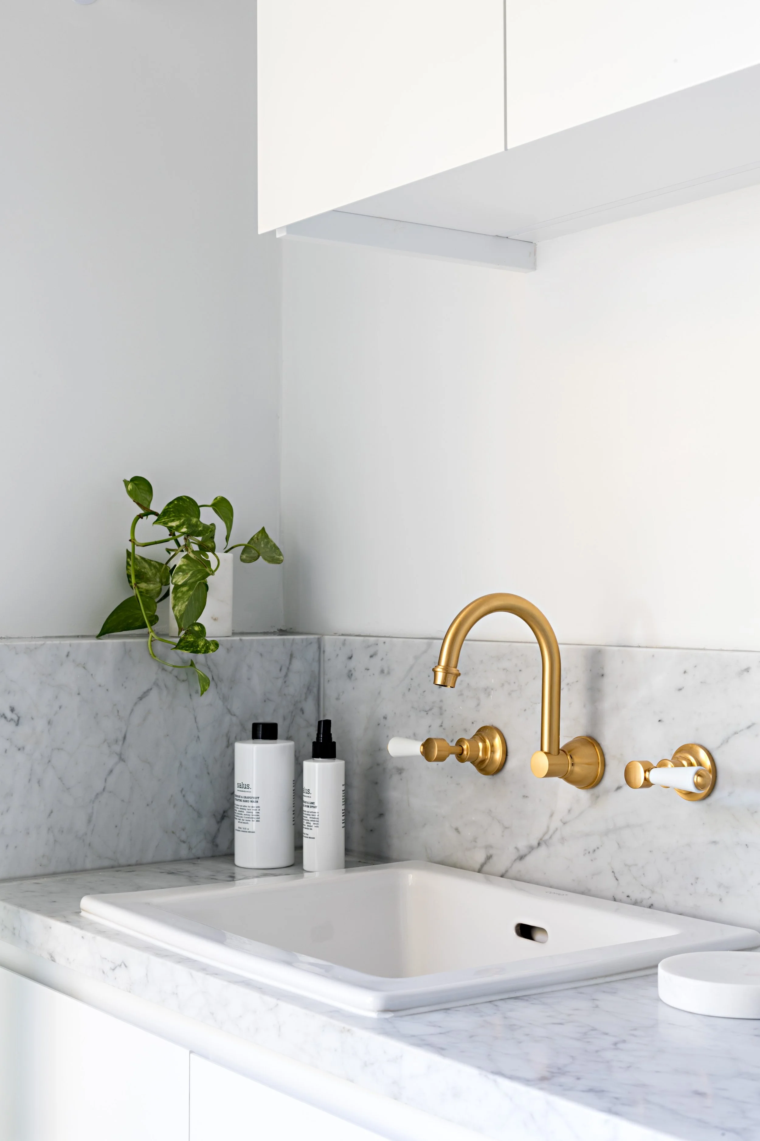 Laundry sink with marble benchtop and splashback, white cabinetry and brass wall-mounted tapware.