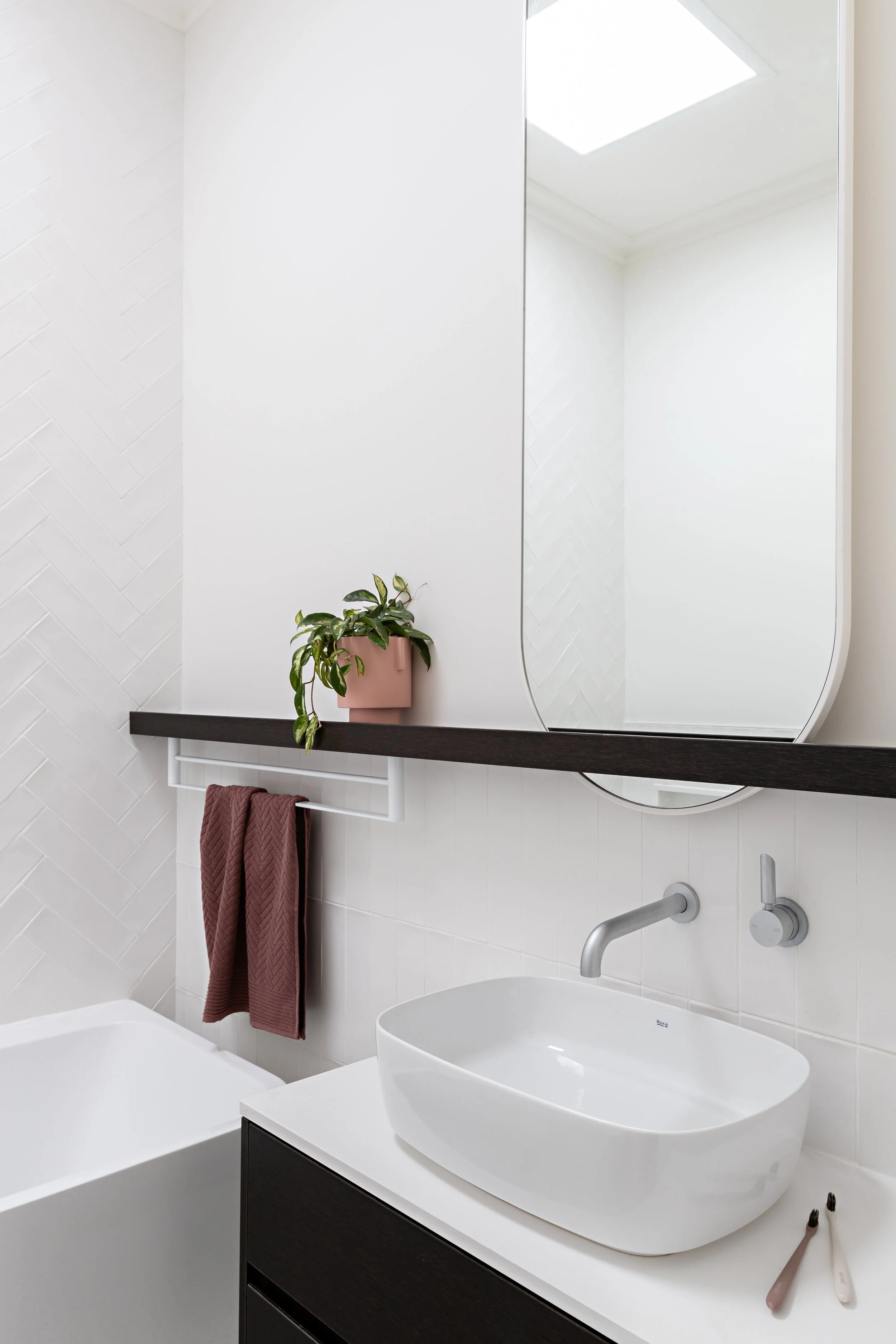 Bathroom vanity with dark timber cabinetry, oval mirror and wall-mounted tapware at Brighton East House.
