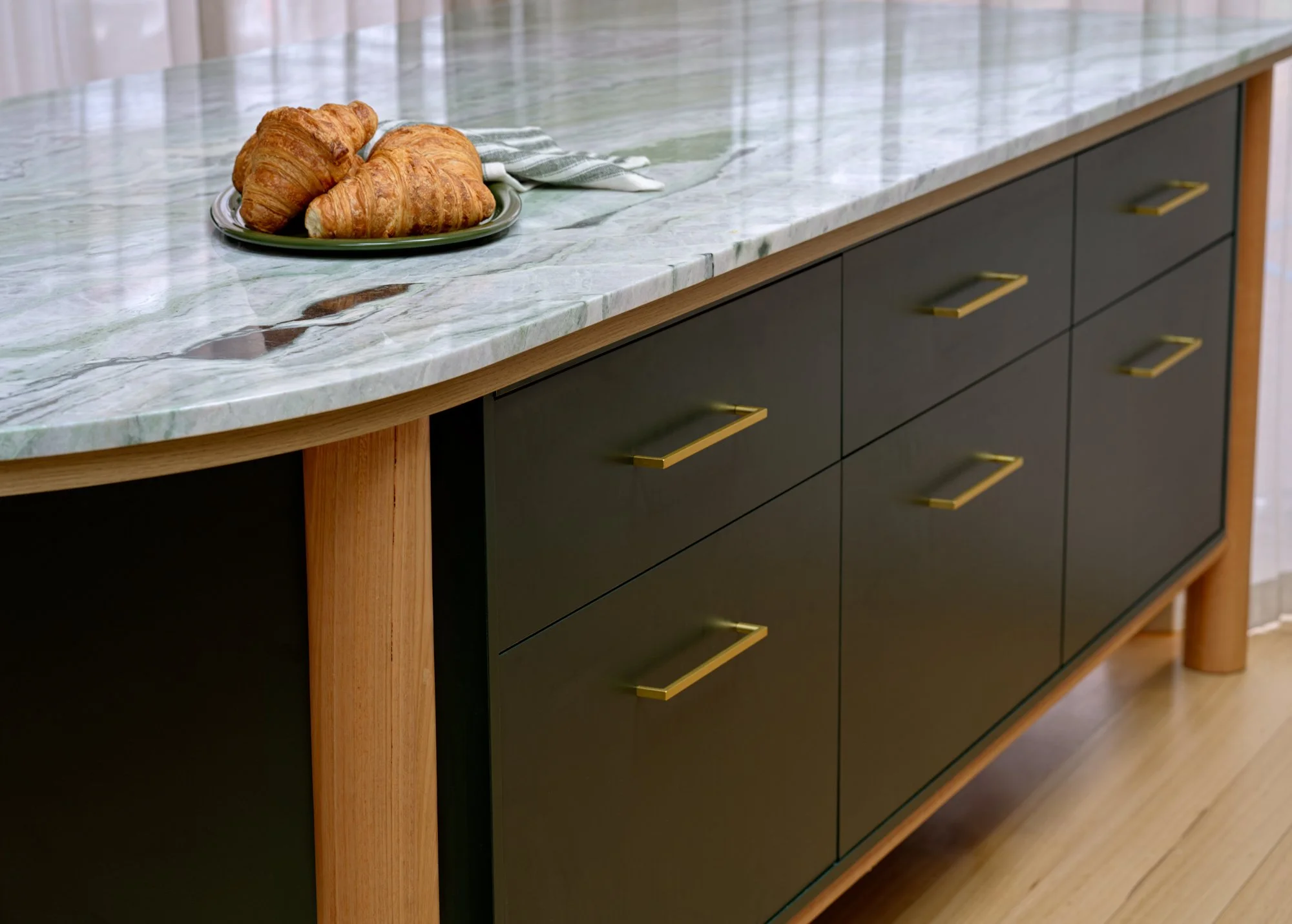 Detail of the Northcote Extension kitchen island with green cabinetry, brass handles, timber framing and a curved stone benchtop.