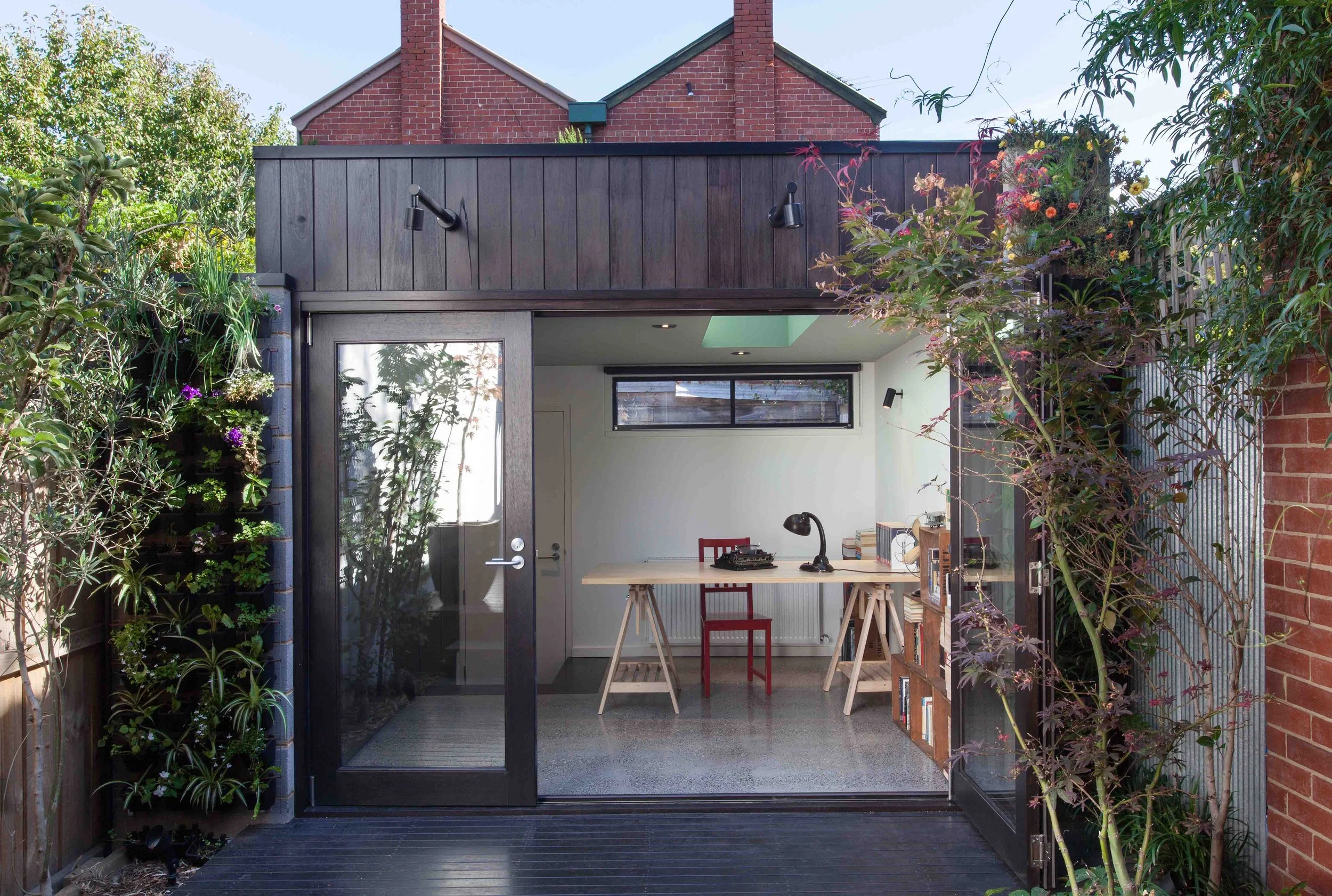 Garden studio with dark timber cladding, glazed entry and large opening connecting the interior workspace to the courtyard.