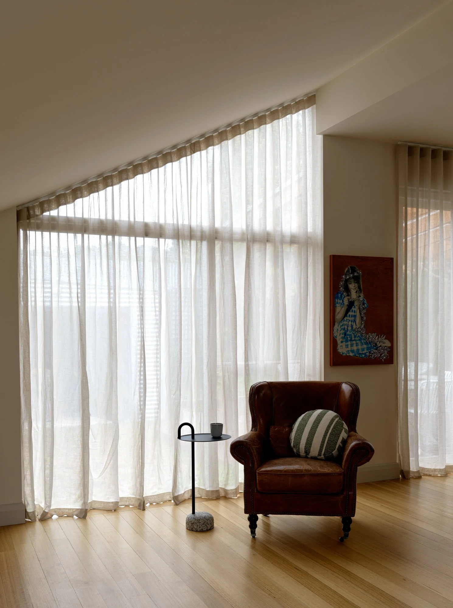 Light-filled living area at Northcote Extension with sheer curtains, timber flooring and a leather armchair.