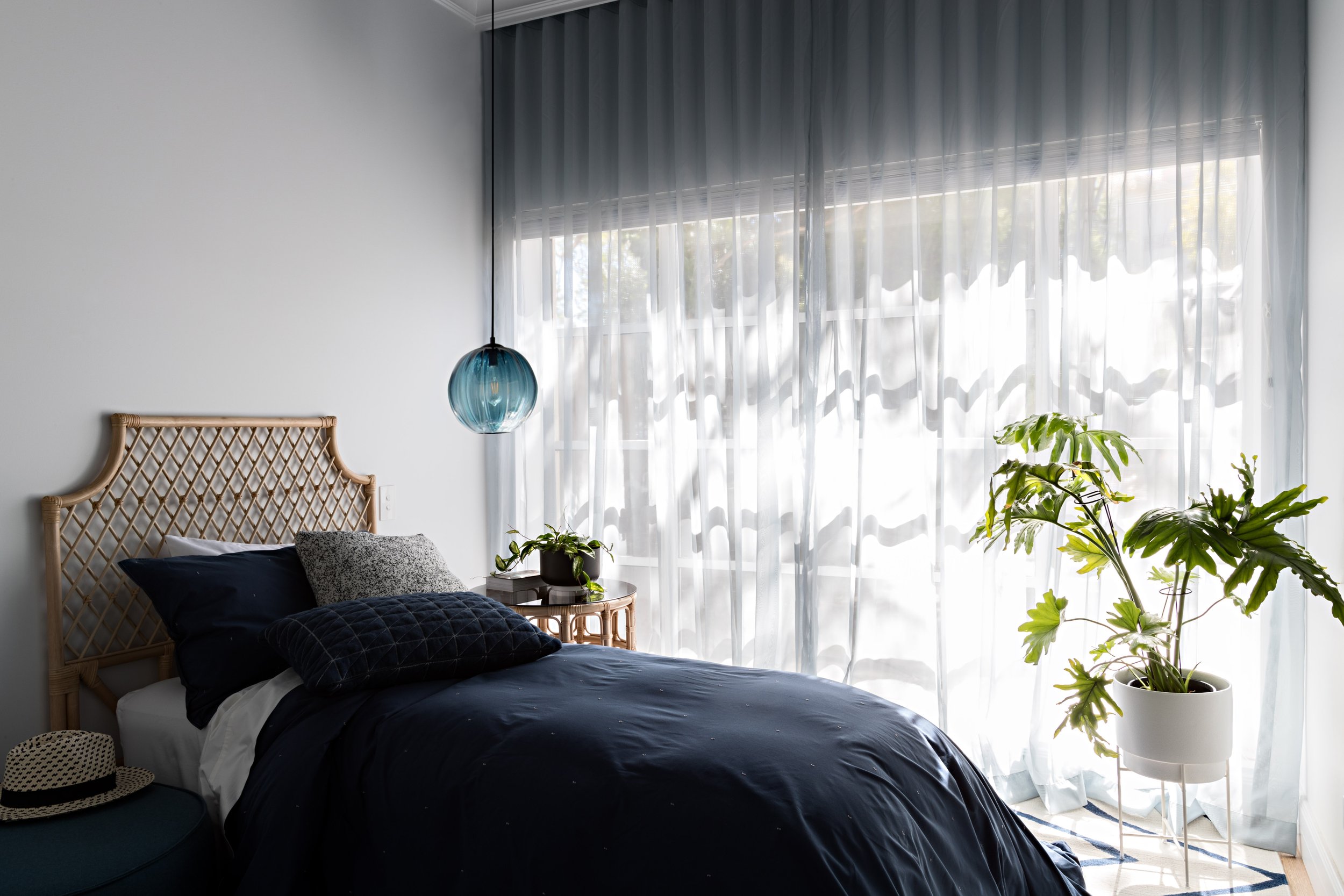 Light-filled bedroom with woven bedhead, blue pendant light and floor-to-ceiling sheer curtains.