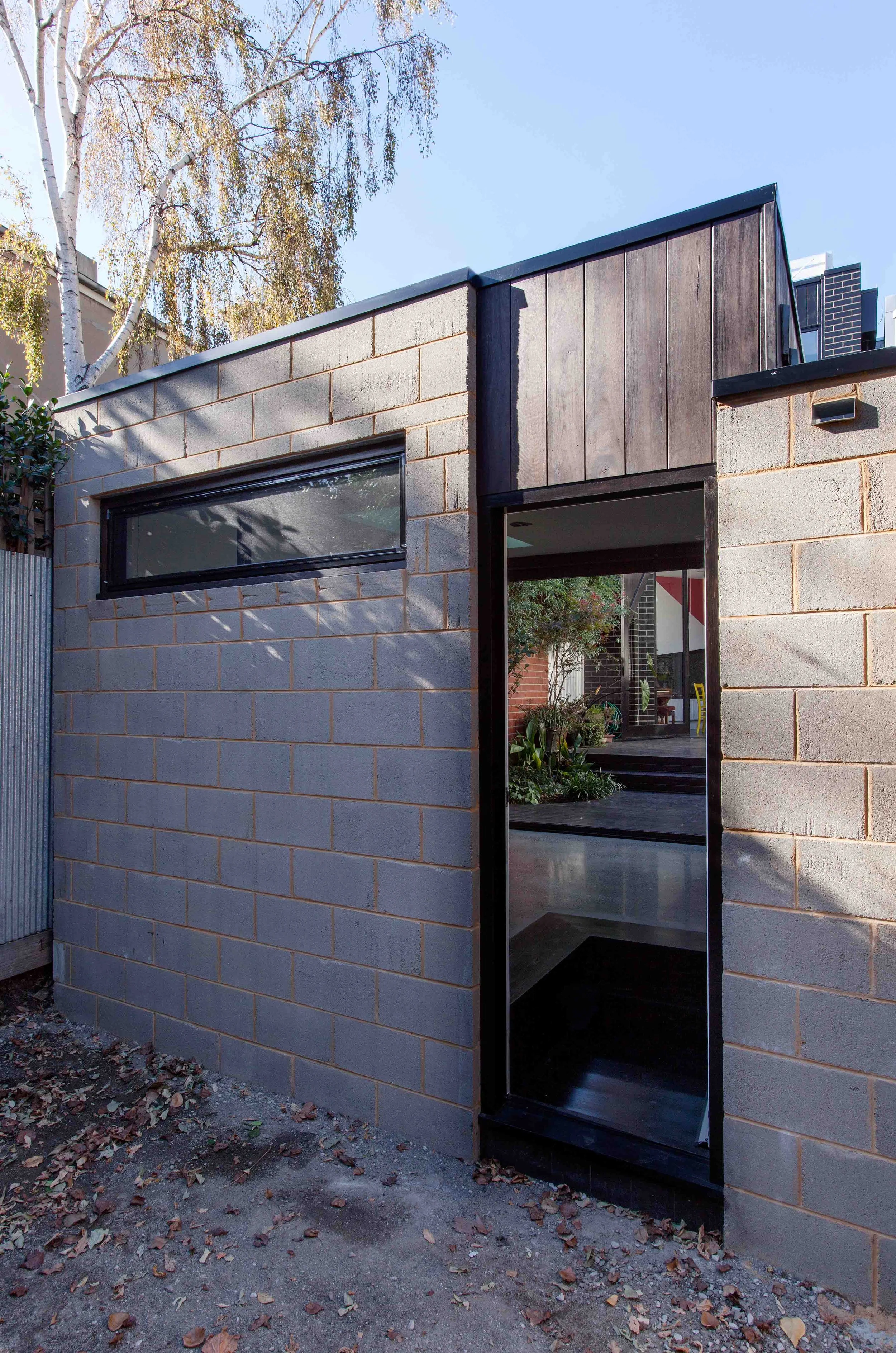 Rear exterior of a compact garden studio combining grey masonry walls, dark timber cladding and a horizontal window.