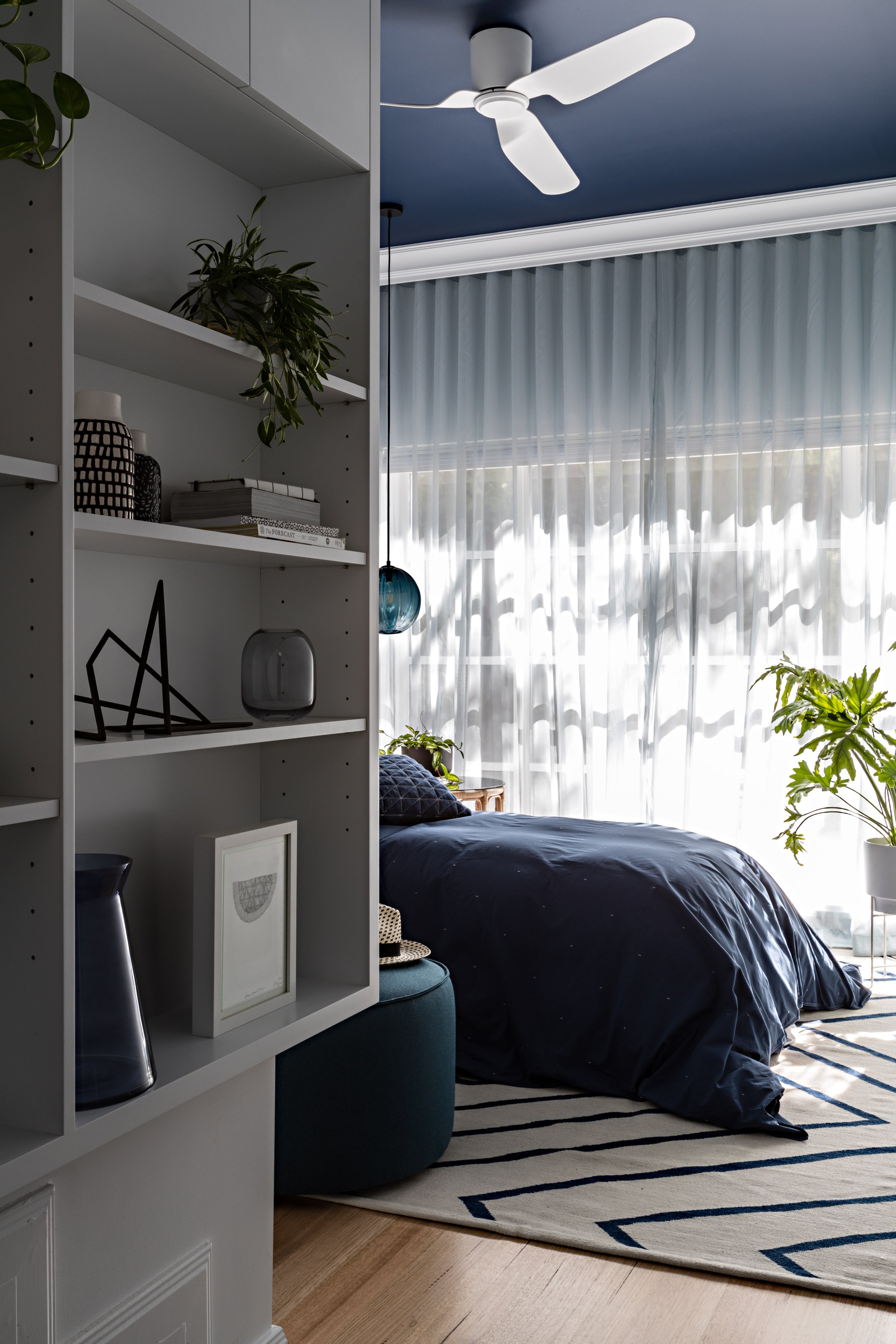 Bedroom with built-in open shelving, dark blue ceiling and floor-to-ceiling sheer curtains.