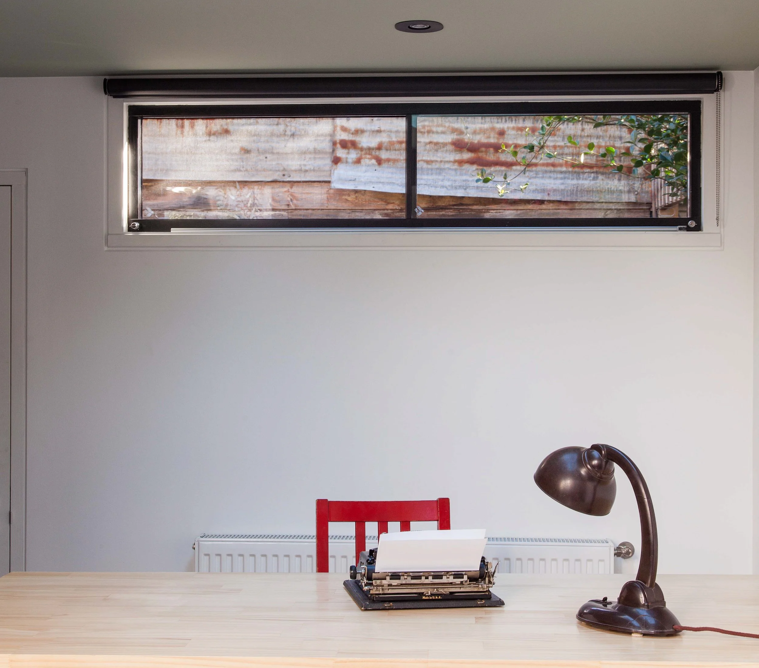 Minimal studio workspace with a timber desk, vintage typewriter, red chair, desk lamp and horizontal garden window.