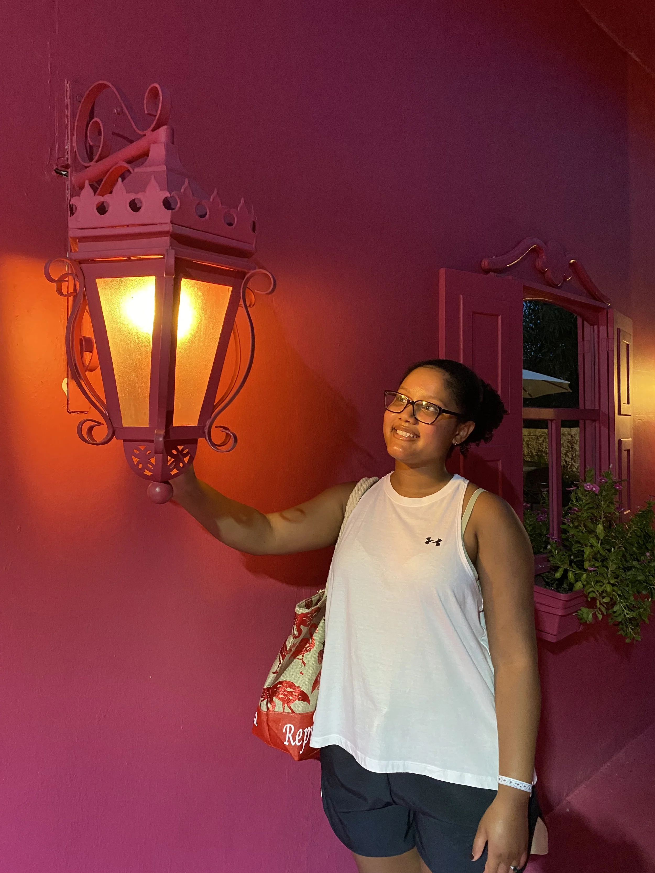Person standing next to a decorative pink wall lamp on a pink wall, wearing a white sleeveless shirt and glasses, smiling.