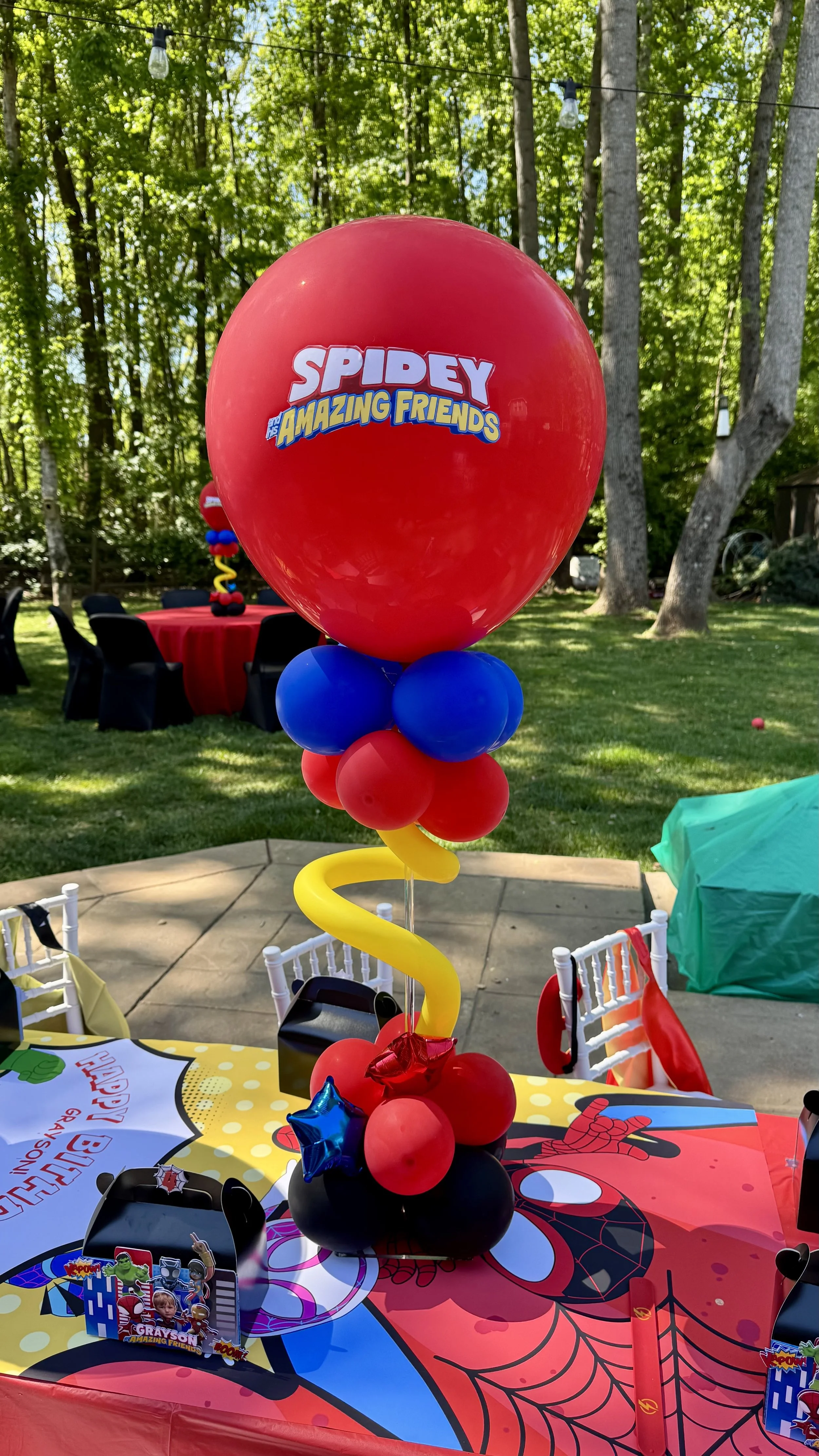 Colorful balloon centerpiece with 'Spidey and His Amazing Friends' logo at a children's birthday party outdoors, surrounded by themed decorations and table settings.
