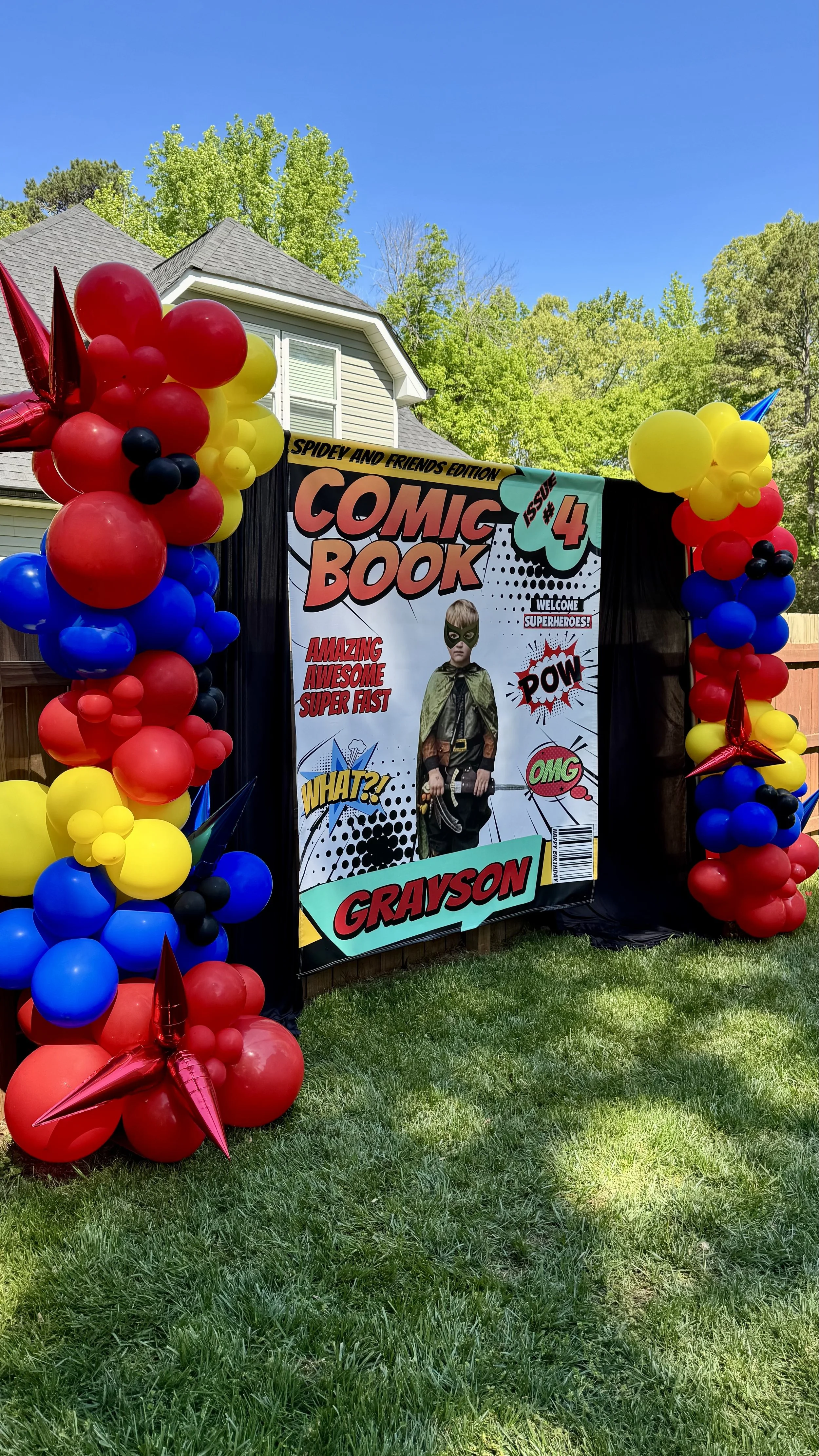 Colorful balloon arch with red, yellow, blue, and black balloons surrounds a comic book-themed backdrop featuring a child dressed as a superhero, with phrases like "Amazing," "Super Fast," "Pow," and "OMG." The setting appears to be an outdoor party 
