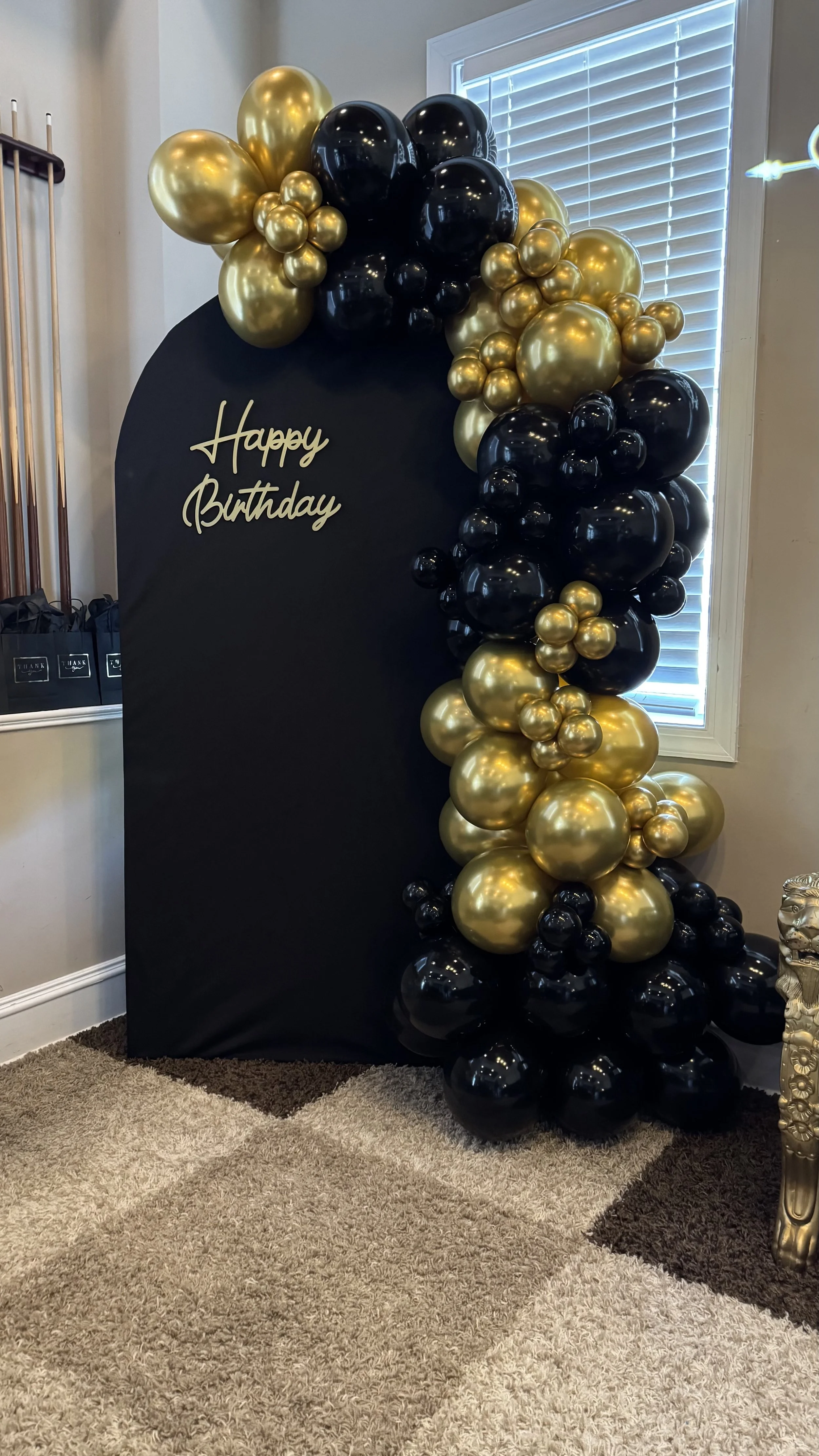 Black backdrop with gold and black balloons arranged around it, with a gold 'Happy Birthday' sign on the backdrop, near a window with white blinds.