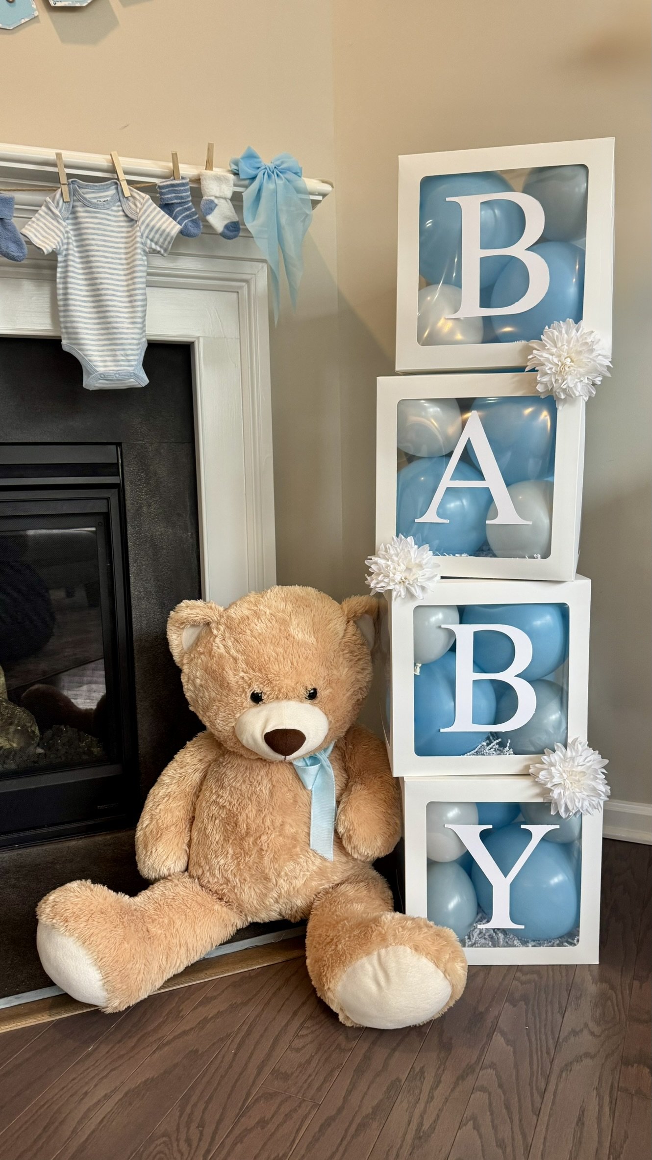 Decorative setup for a baby boy's nursery with a large teddy bear, stacking cubes with blue balloons and white letters spelling B-A-B-Y, a clothing hanging on the mantel, and white and blue accents.