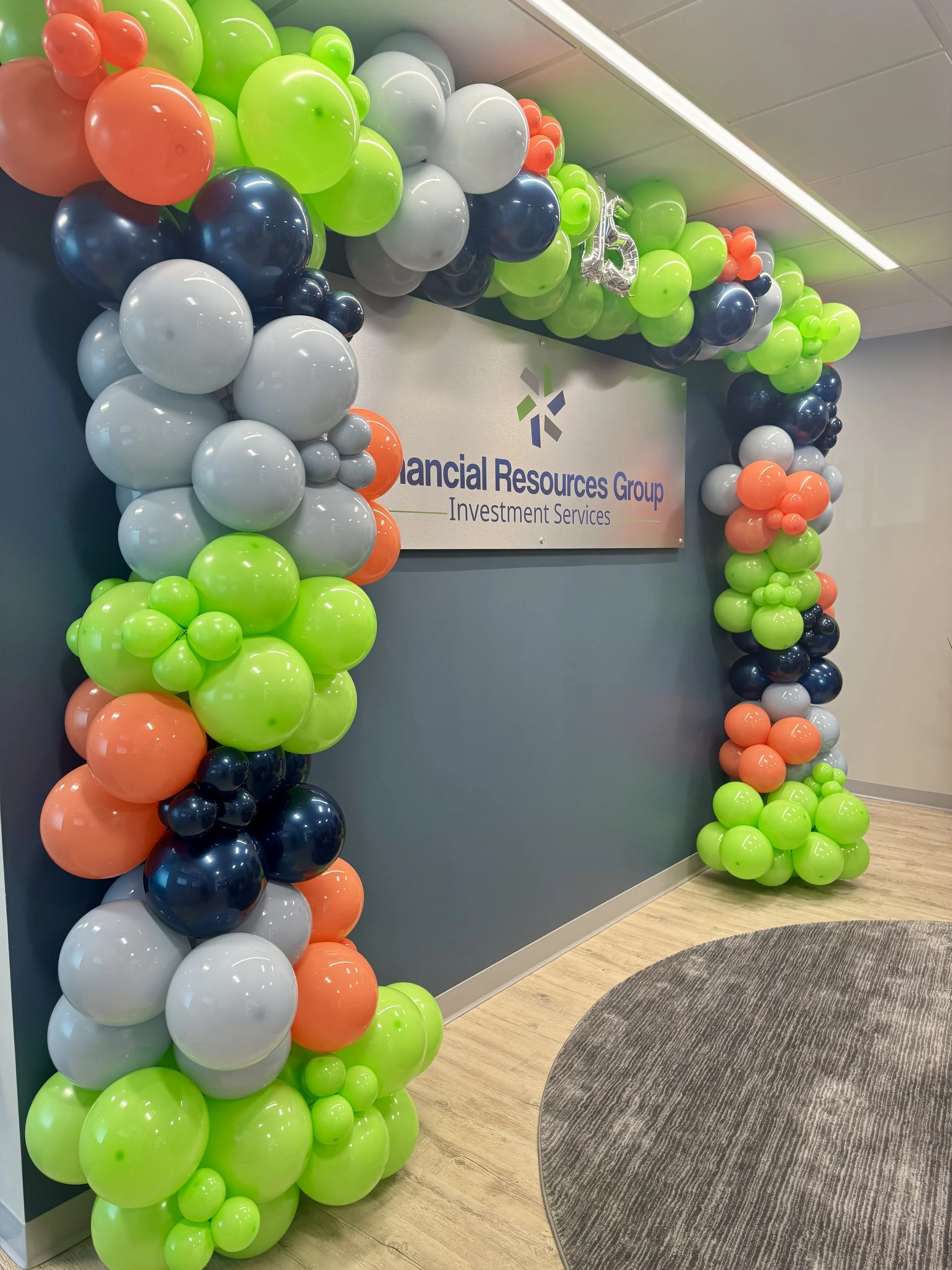 Decorative balloon arch featuring green, gray, coral, and navy balloons framing a sign inside an office.