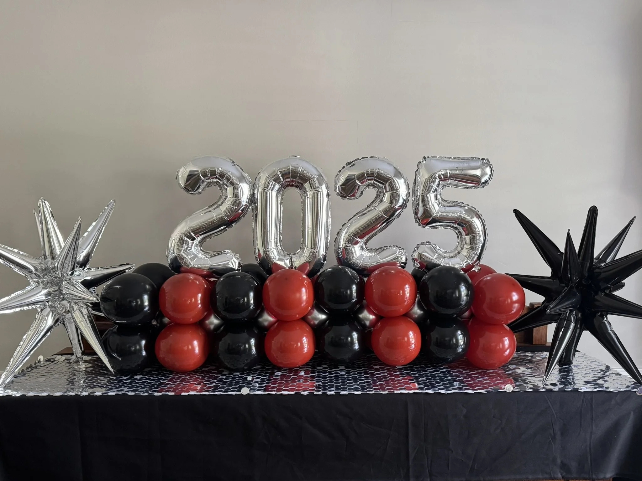 Decorative table with silver and black balloons, red balloons, and large metallic balloons spelling out '2025', flanked by silver and black star-shaped balloons.