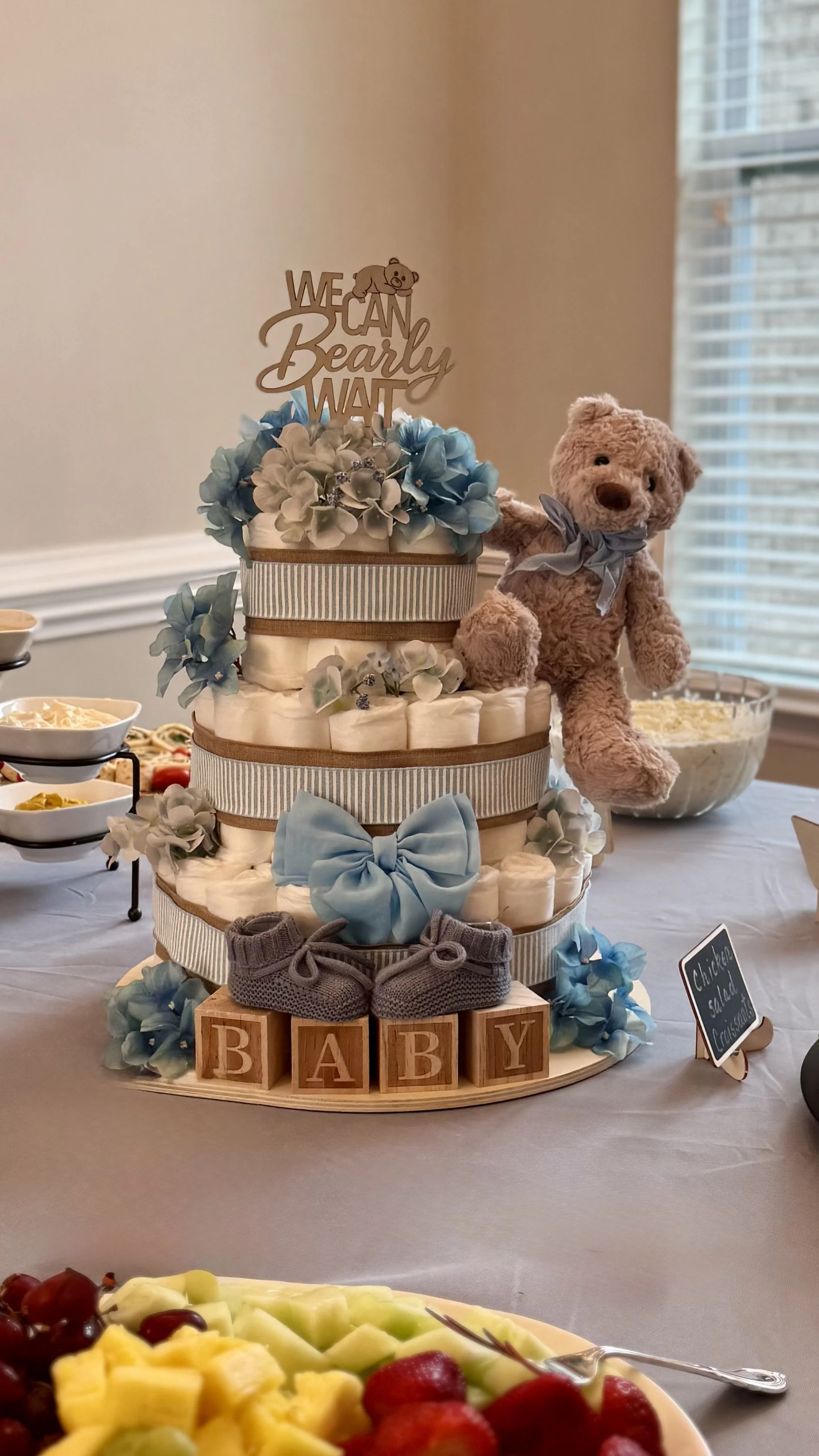 A baby shower cake decorated with blue ribbons, flowers, a teddy bear, and a cake topper that says 'We Can Bearly Wait.' Wooden blocks spell out 'BABY' at the base of the cake. A plush teddy bear is placed next to the cake.