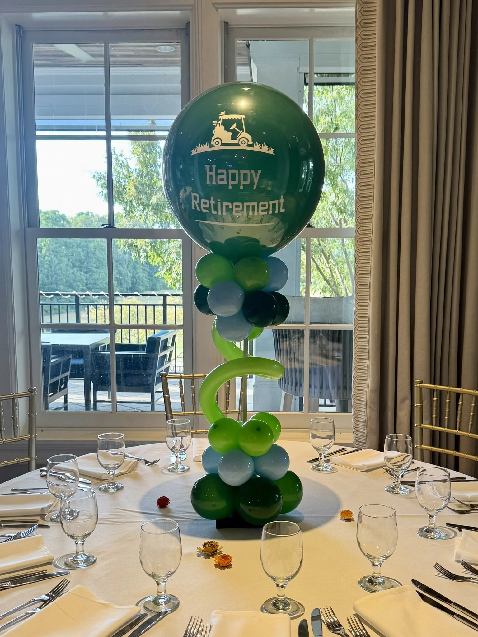 Decorative centerpiece with a large green balloon reading 'Happy Retirement', surrounded by green, white, and black balloons, arranged on a banquet table inside a room with large windows overlooking an outdoor patio.