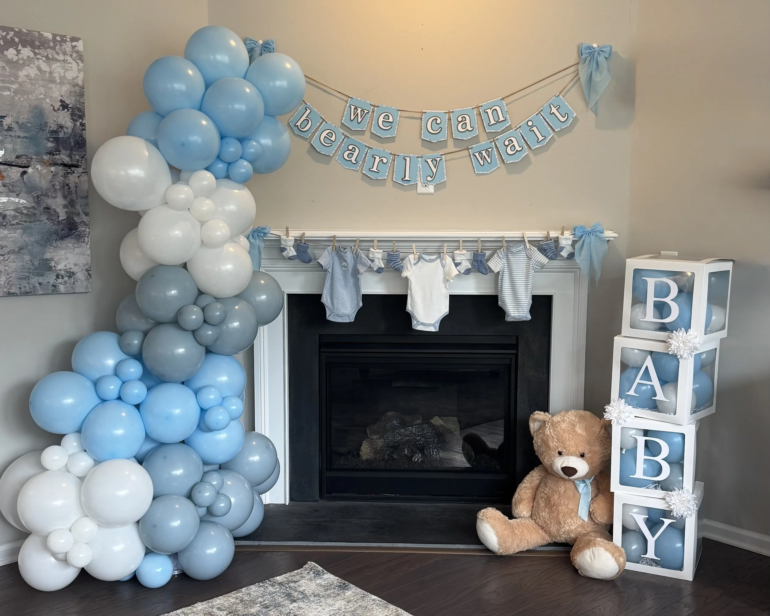 Baby shower decorations with blue and white balloons shaped in a column to the left of a fireplace. Above the fireplace hangs a banner with blue background and white letters spelling 'we can bearly wait.' Small baby clothes are hanging on a line above the fireplace, and a large teddy bear with a blue ribbon sits on the floor next to a white block with stacked cubes spelling 'BABY' in blue letters.