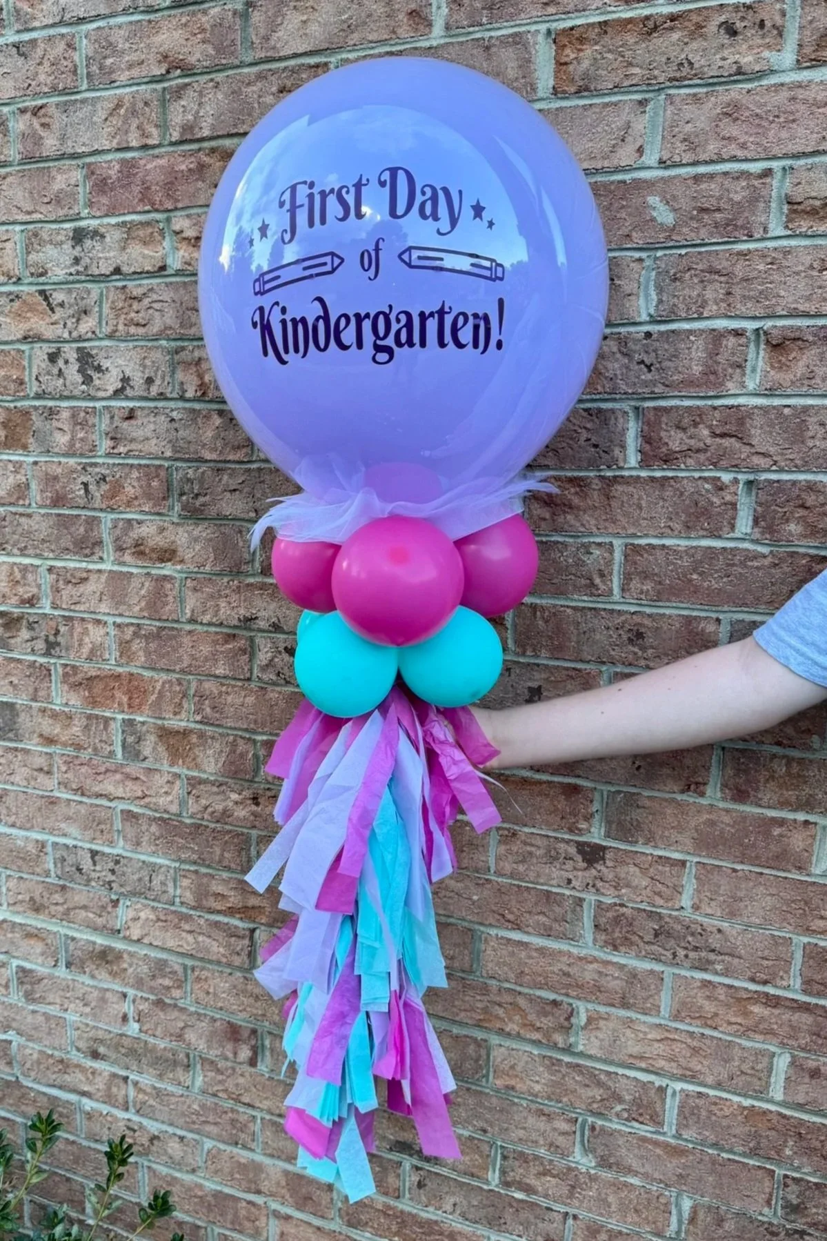 A back to school balloon wand display with a large purple balloon inscribed with 'First Day of Kindergarten!', decorated with smaller pink and turquoise balloons, ribbons, and tissue paper streamers.