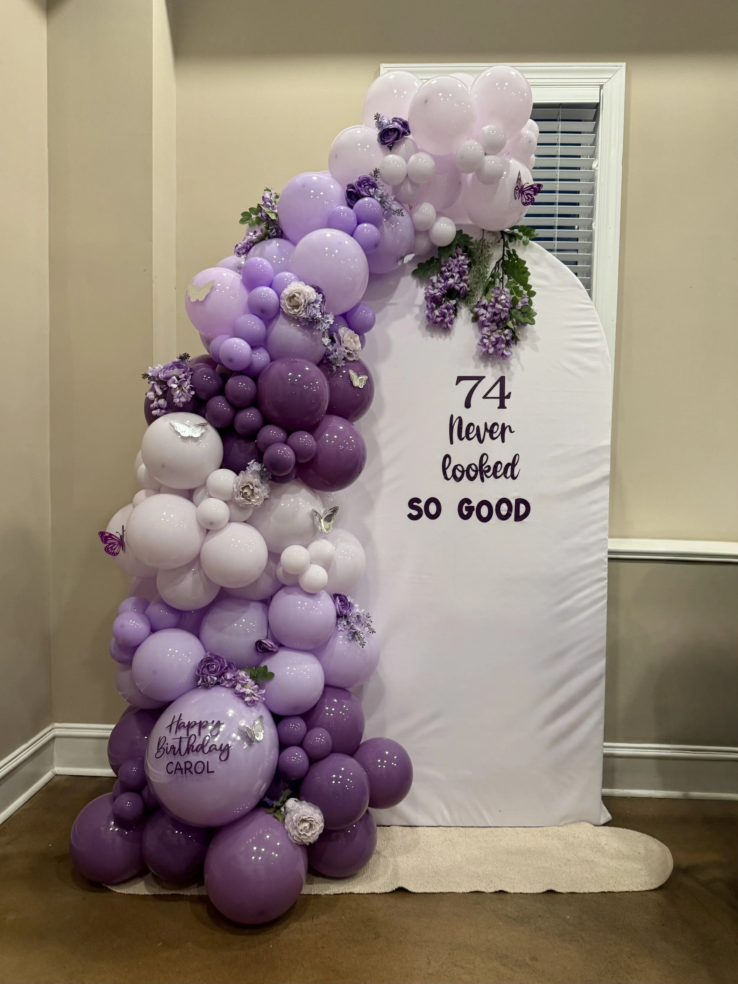 Elegant purple balloon arch with colors ranging from lavender to deep plum, accented with florals, greenery, and butterfly details. This custom balloon backdrop includes a personalized birthday balloon, creating a stylish centerpiece for milestone bi