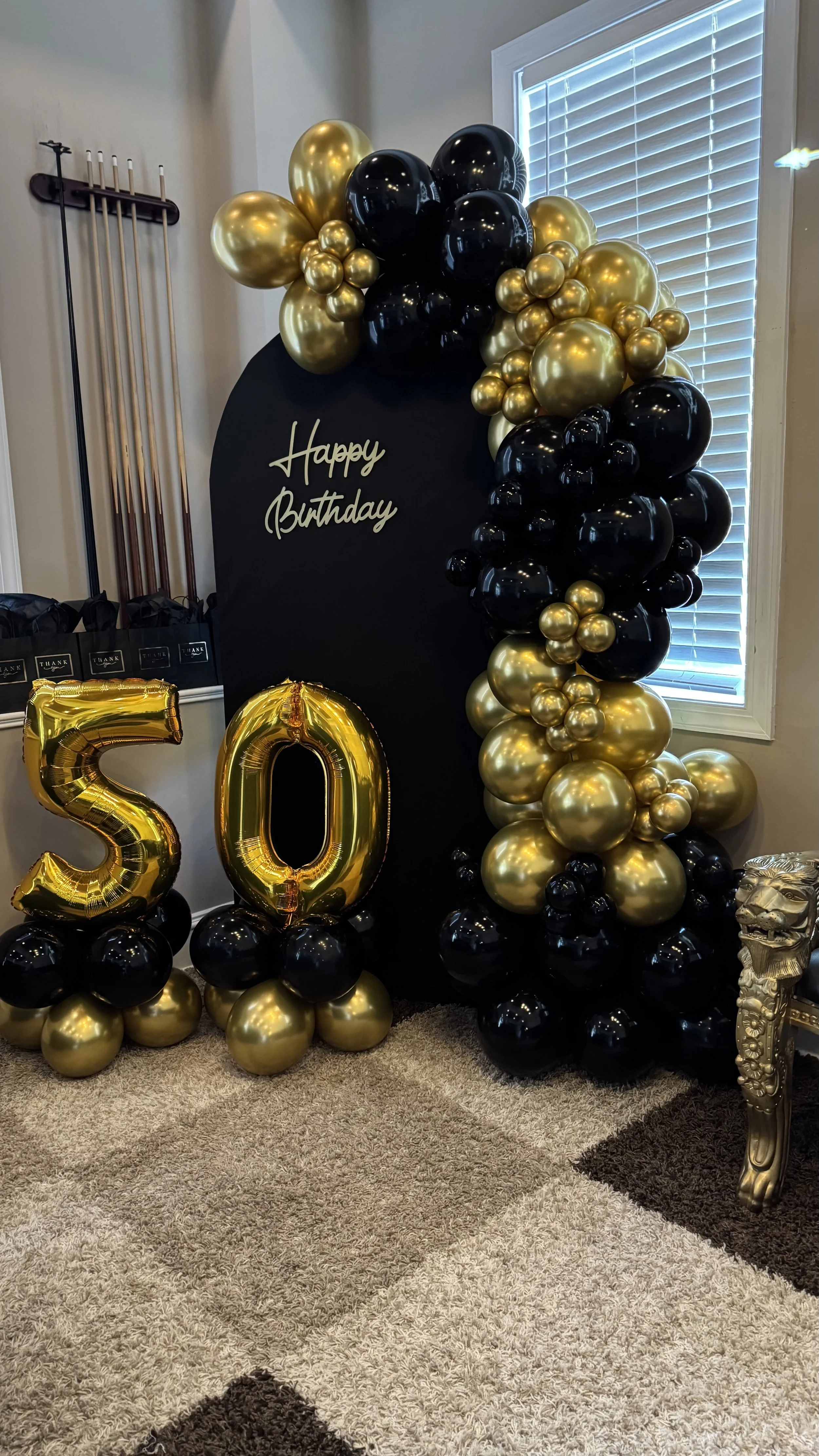 Decorative birthday display with black, gold, and black balloons, a black backdrop with "Happy Birthday" written in white, and large gold number balloons forming '50'. The setup is indoors next to a window with blinds and a gold decorative side table